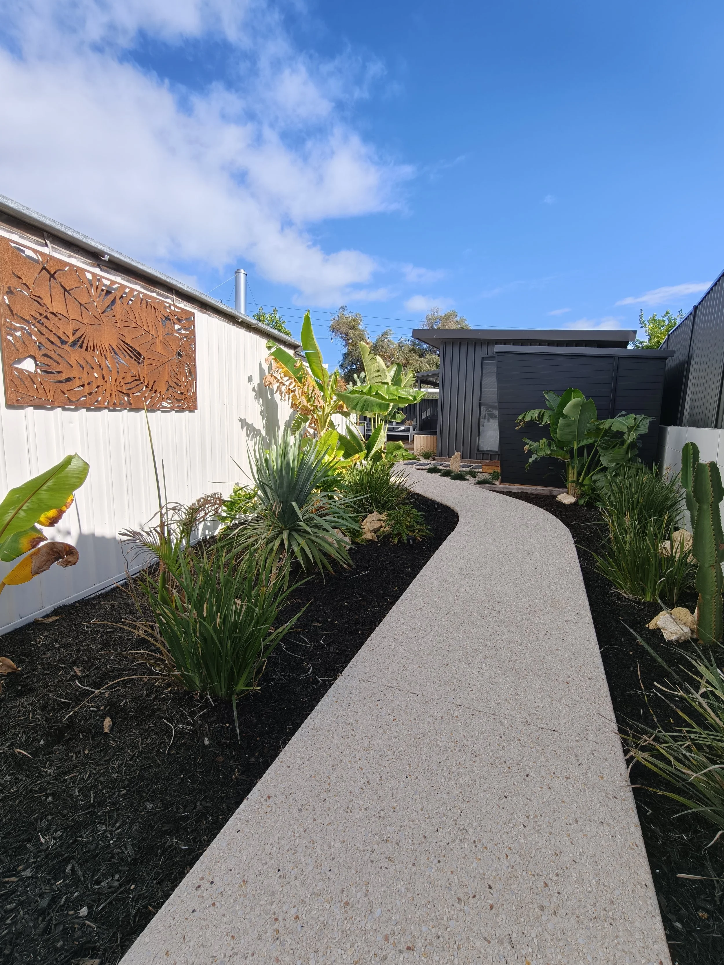 A garden pathway curves through a landscaped yard with plants and trees, enclosed by a white fence on the left and dark-colored structures on the right, under a blue sky with some clouds.