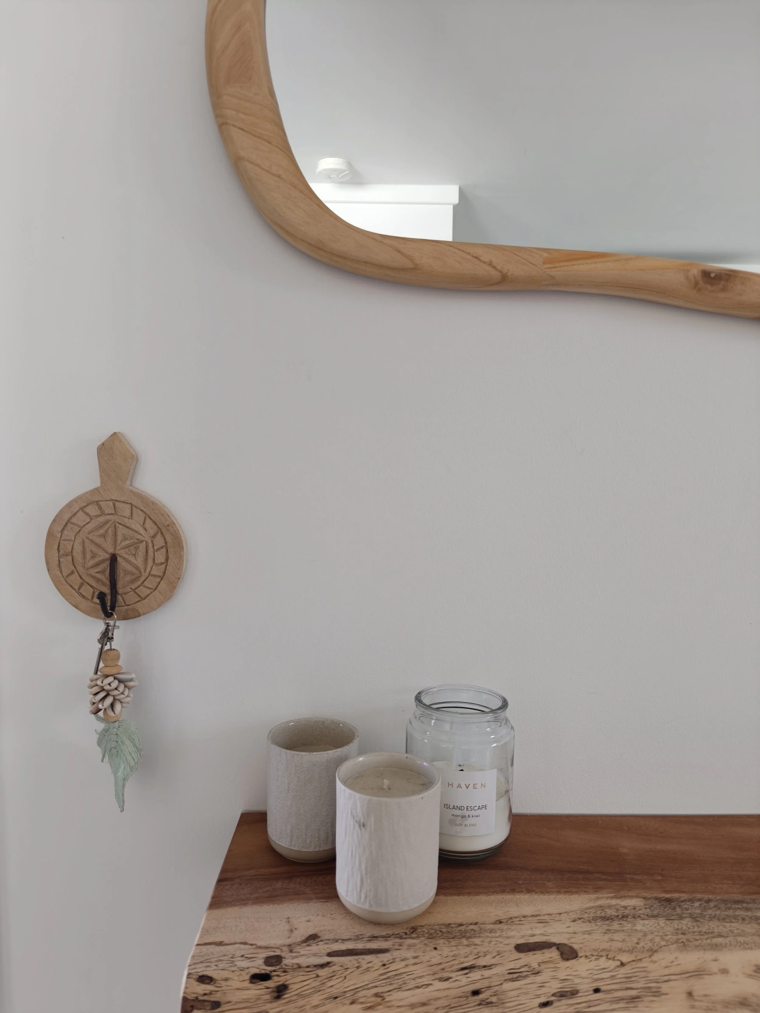 A wooden tray displays candles and a glass jar on a wooden surface against a white wall, with a mirror, a decorative wall hanging, and a small object on the wall in the background.