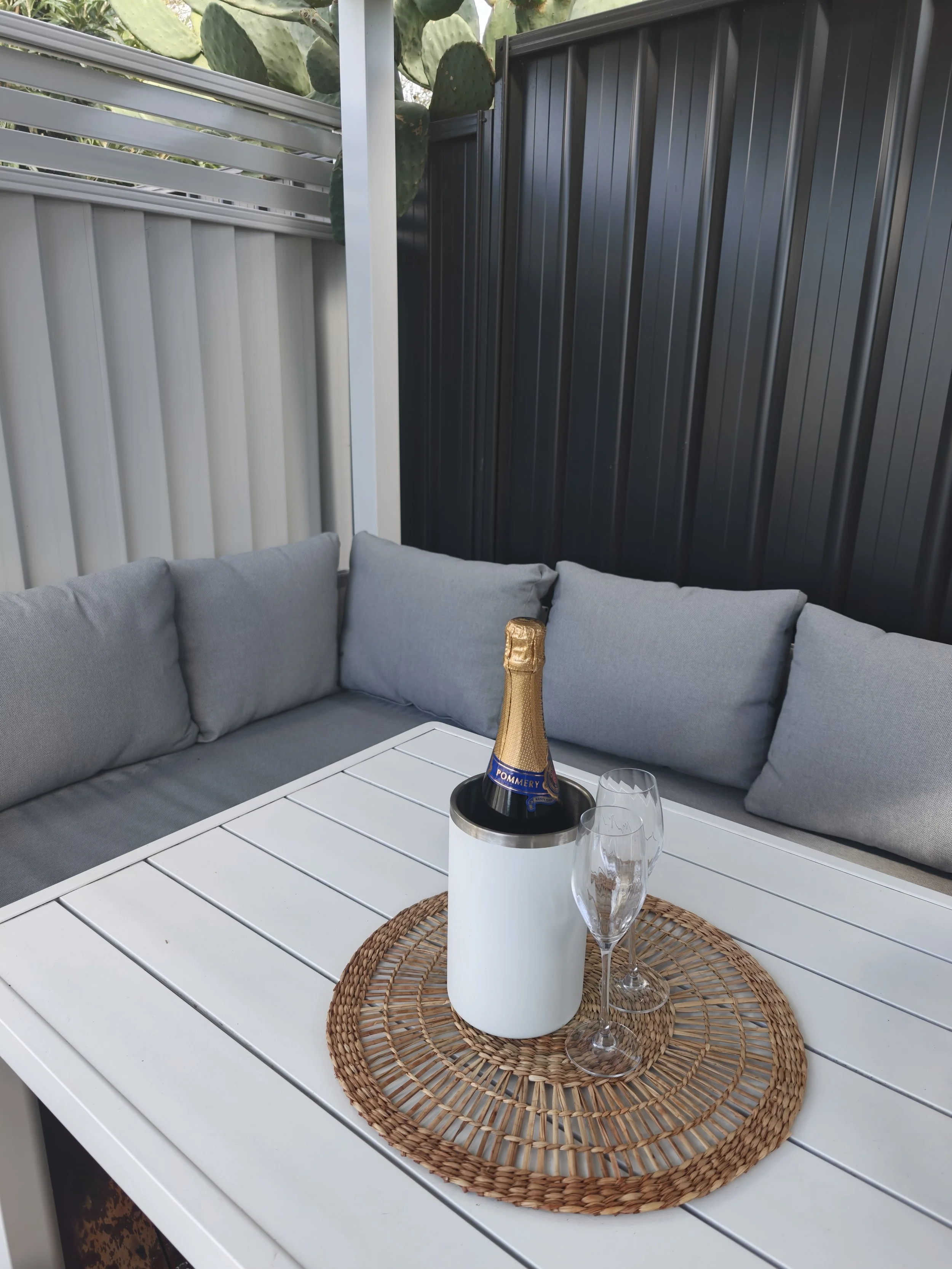 A white outdoor table with a woven placemat, a white ice bucket with champagne, a champagne bottle, and two empty champagne glasses, set against a background of gray and white cushioned seating and a privacy fence with green cactus plants.