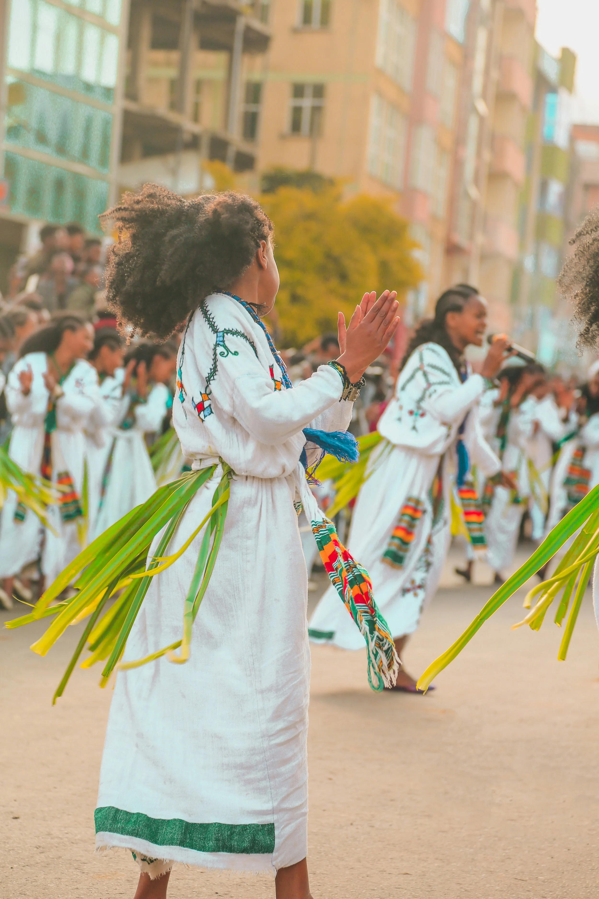 A group of people dressed in traditional white clothing with colorful embroidered details, participating in a cultural outdoor event or festival in an urban area with modern buildings and trees in the background.