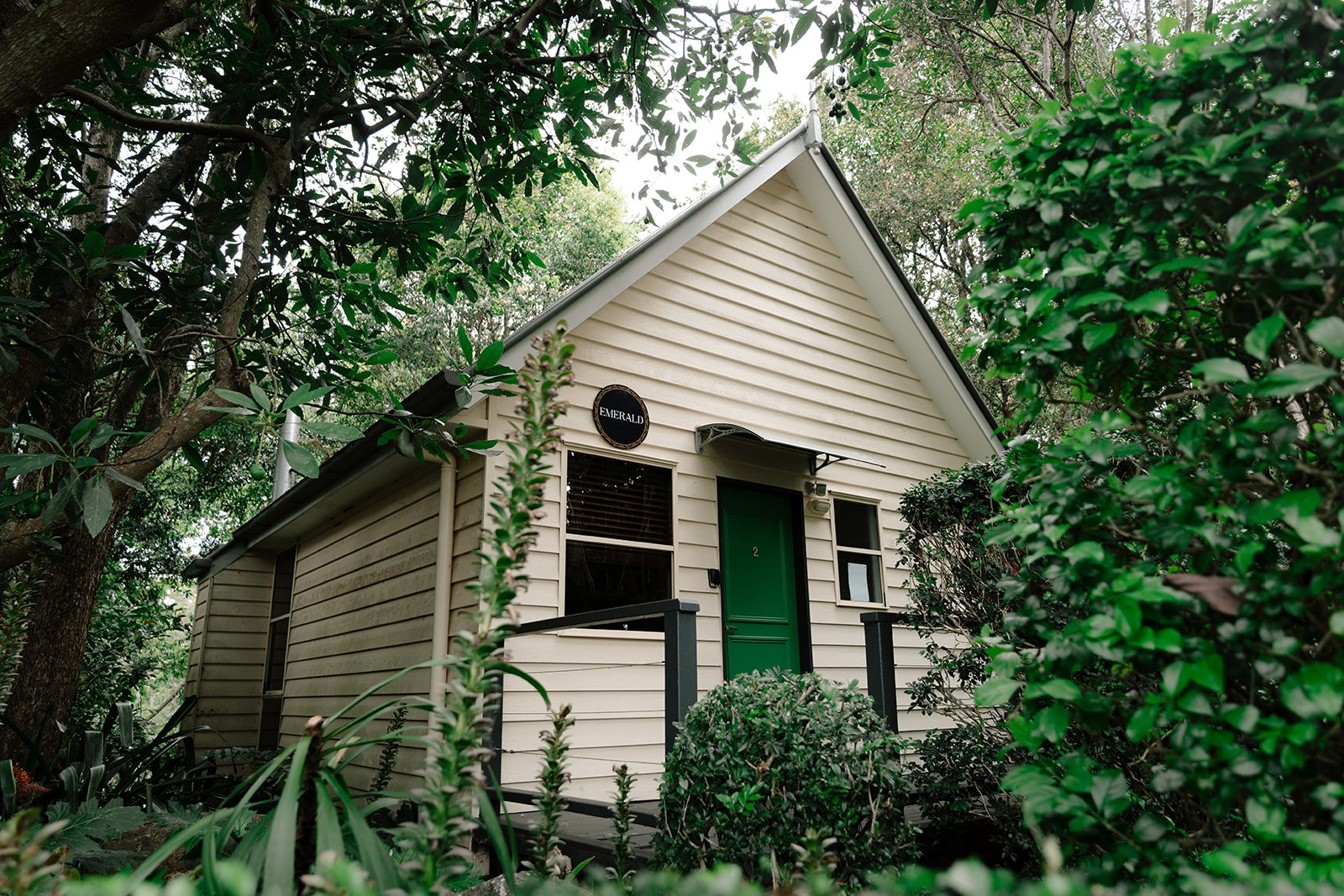 Small house with cream-colored siding, green door, surrounded by lush green trees and bushes.