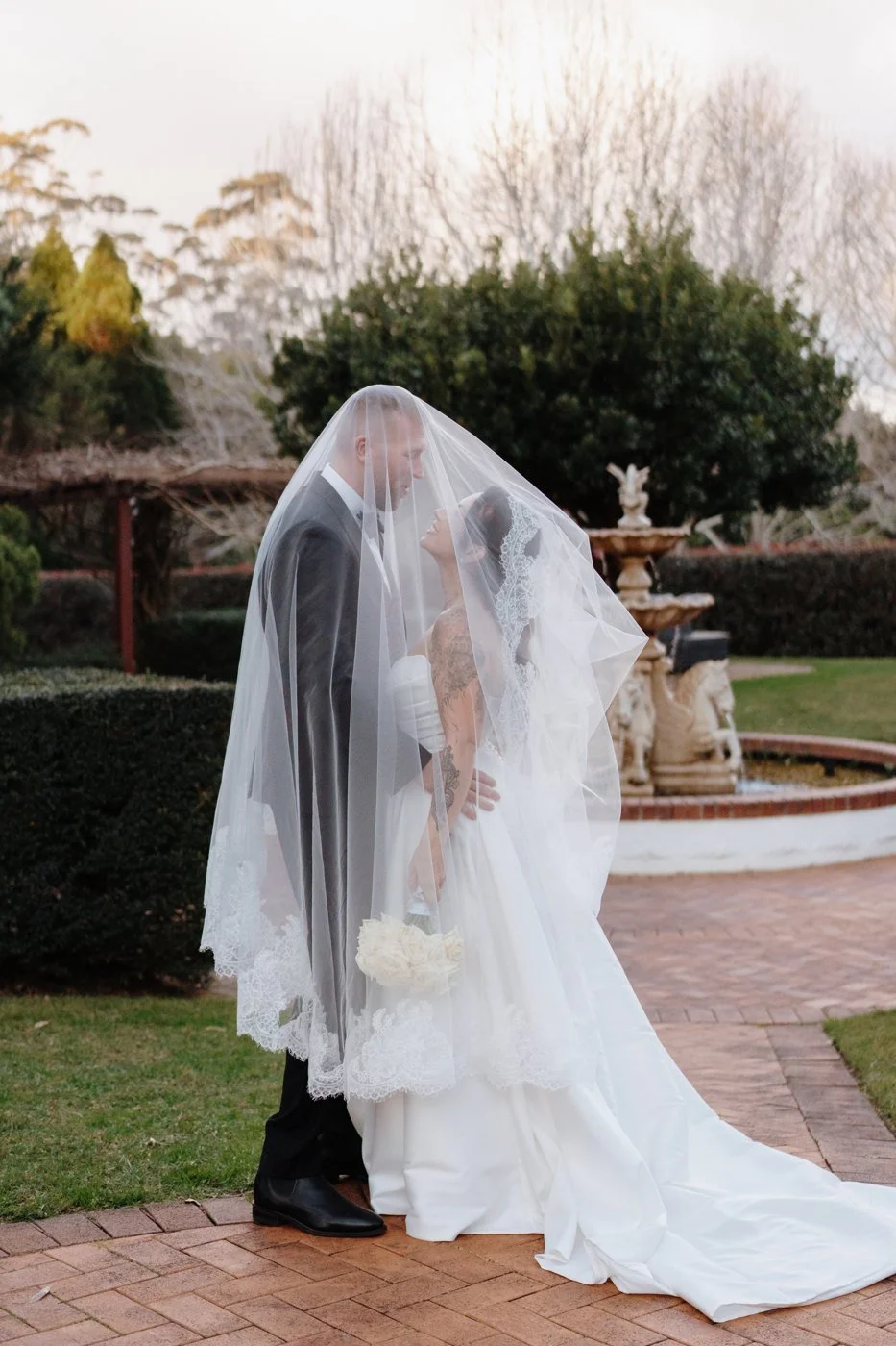 A wedding couple share a kiss beneath a veil beside the garden fountain at Shambala Estate.