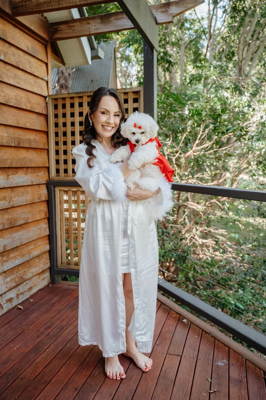 A bride in getting-ready clothes holds a small white dog on a leafy cottage balcony at Shambala Estate.