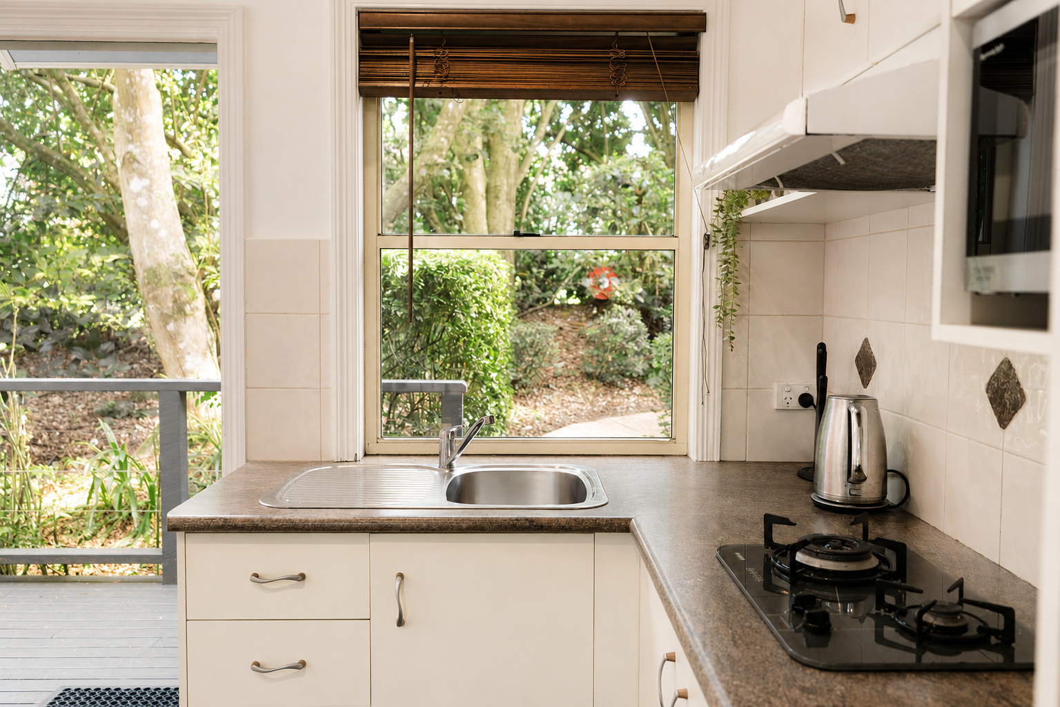 View of a kitchen sink with a window overlooking a garden with trees and greenery. Countertop has a kettle and a stove, with a microwave above.