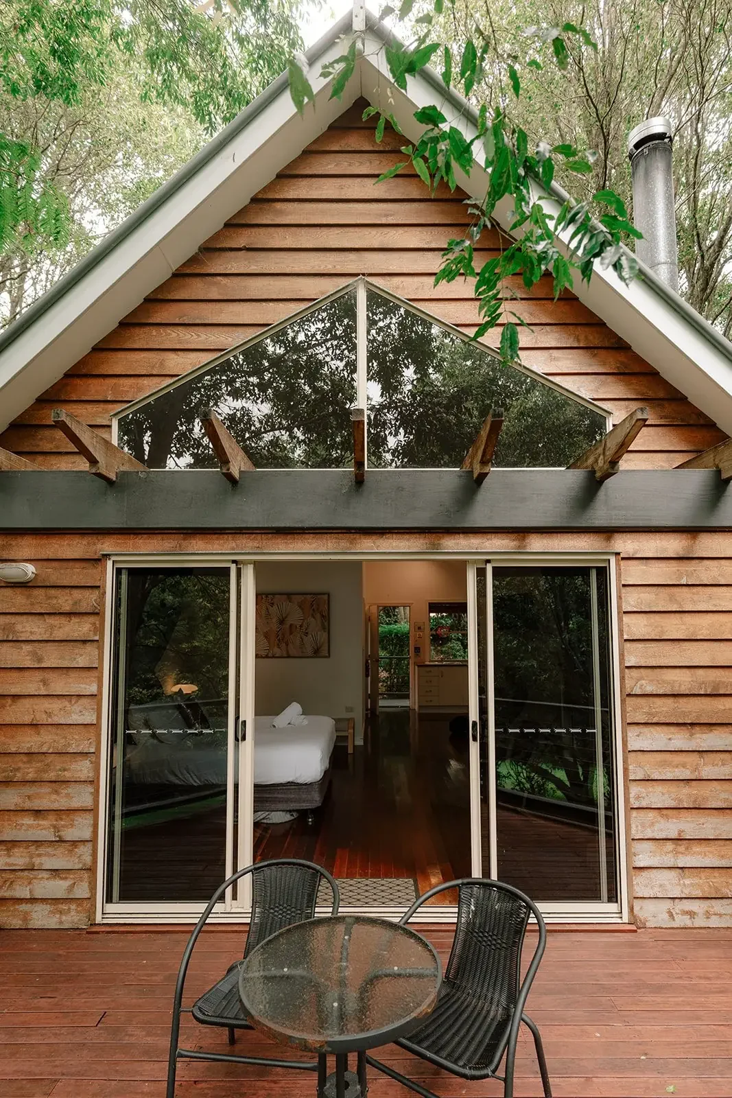 A wooden house with large glass sliding doors leading to a bedroom with white bedding, a small table, and a sliding door in the background, with a round outdoor table and three chairs on the wooden deck outside.