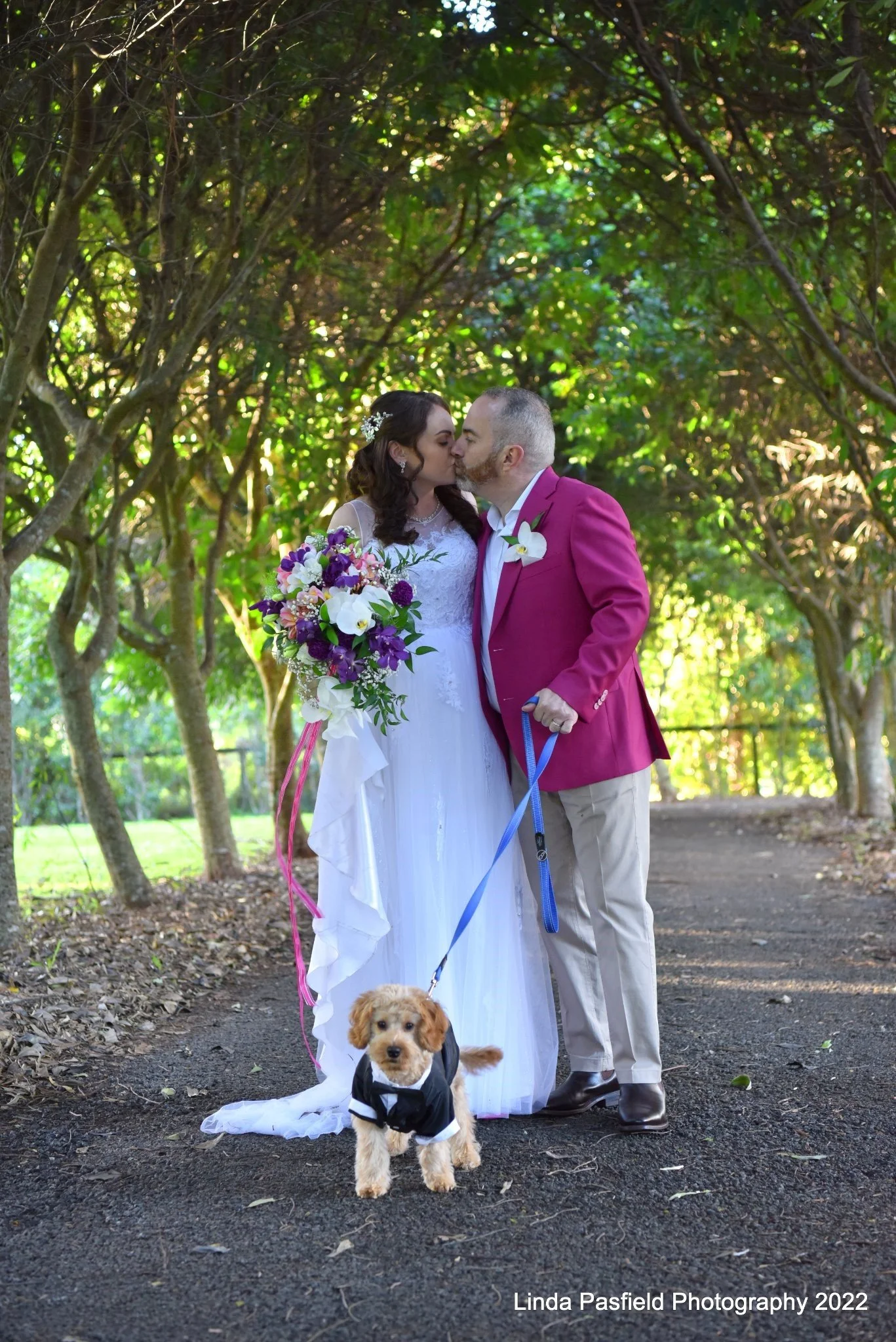 Newlyweds Samantha and Jason share a kiss in the Shambala Estate gardens with their small dog dressed in a tuxedo.