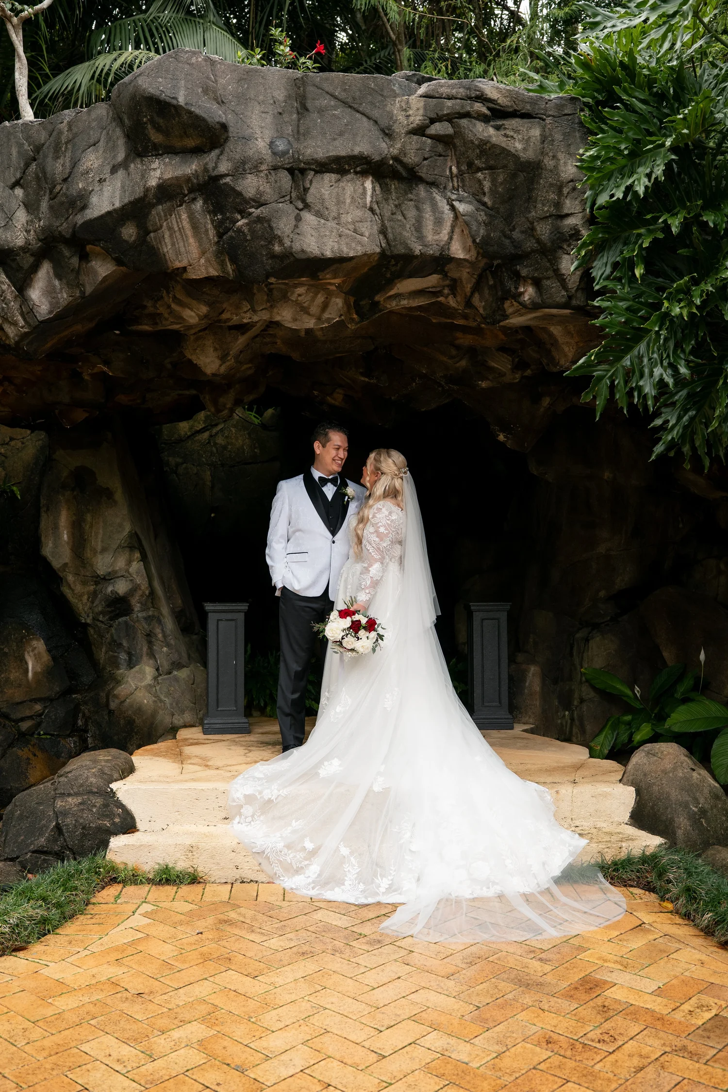 A bride and groom standing together beneath the Waterfall Grotto rock arch at Shambala Estate.