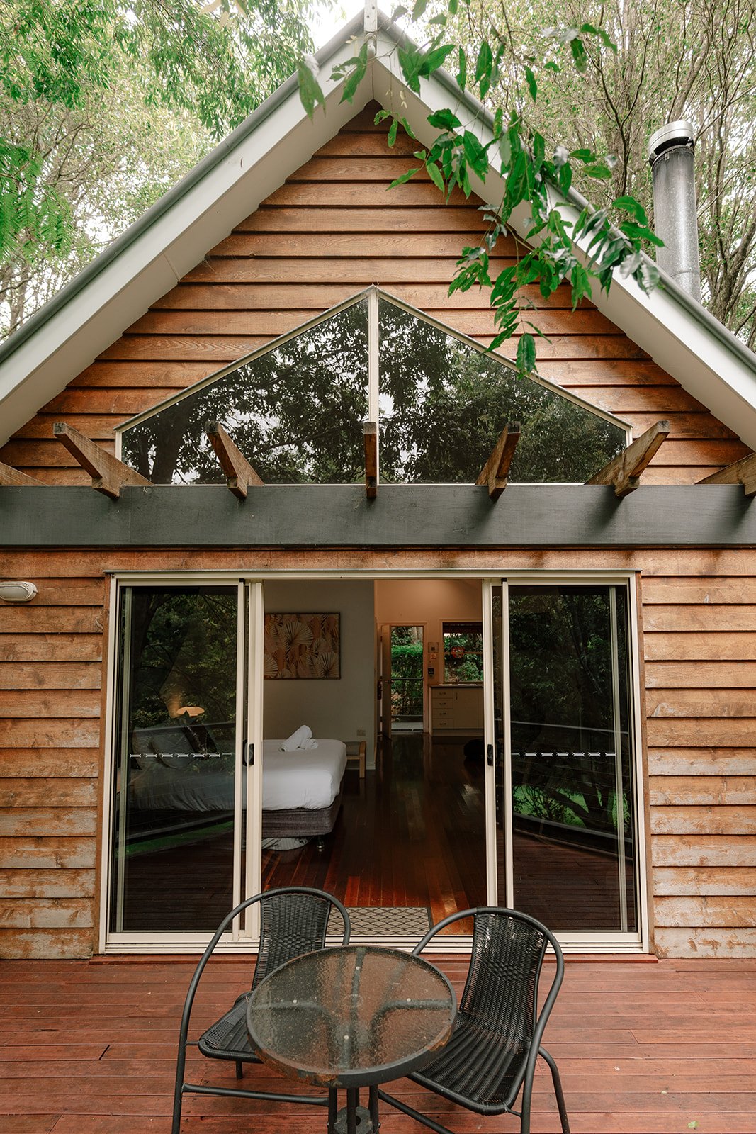 A wooden house with large glass sliding doors leading to a bedroom with white bedding, a small table, and a sliding door in the background, with a round outdoor table and three chairs on the wooden deck outside.