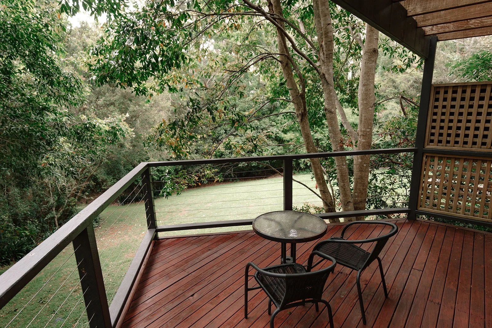 A wooden balcony with a metal table and two chairs, overlooking a lush green backyard with trees and grass.