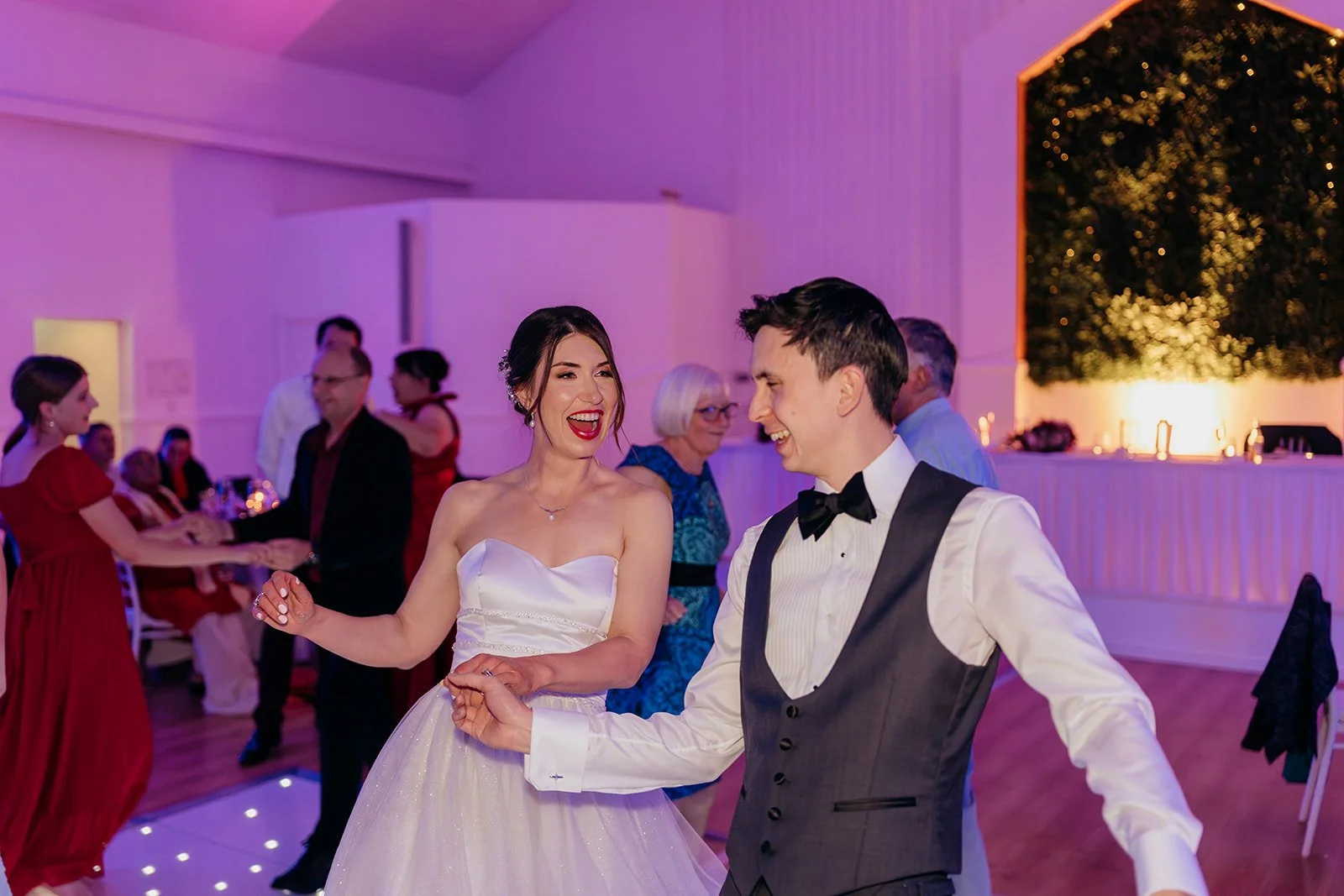 A newlywed couple and guests dancing on the illuminated floor in purple Galaxy Ballroom lighting.