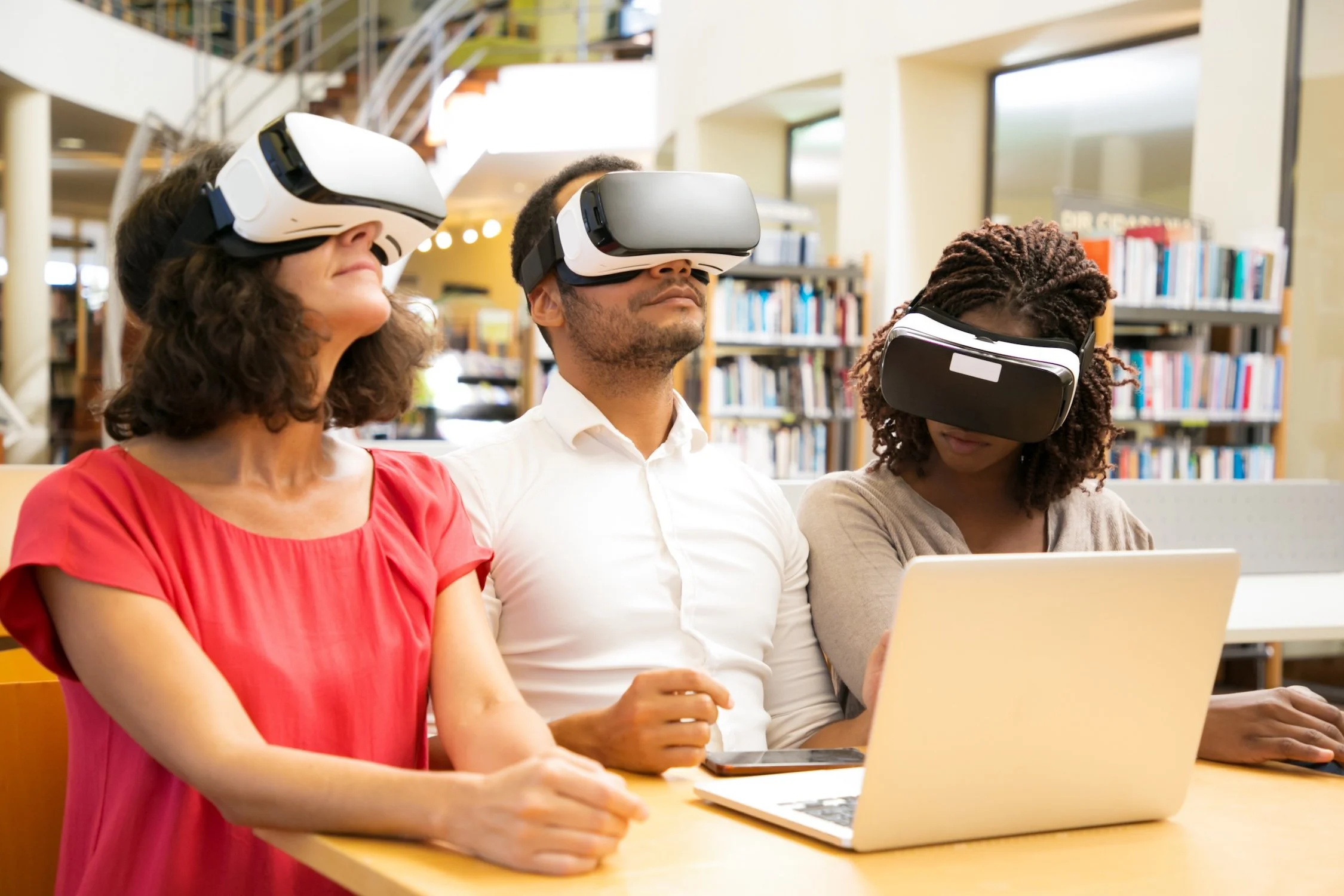 Three diverse people wearing virtual reality headsets sitting at a table with a laptop in a library.