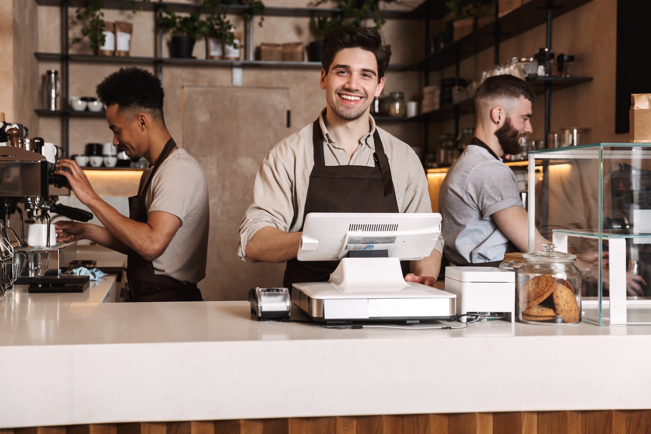Smiling barista working at the counter of a coffee shop with two other baristas in the background.