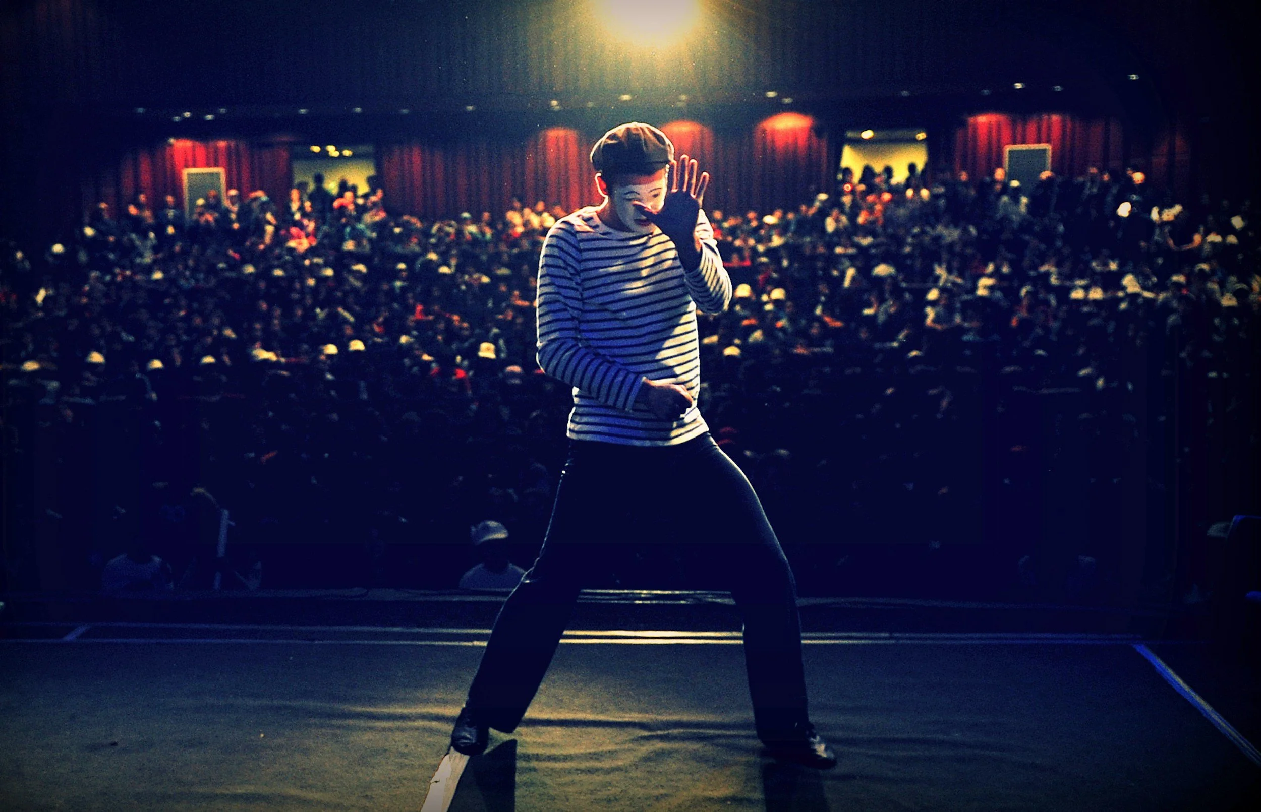 A mime in a striped shirt and black pants performs on a stage in front of a large audience in a theater.