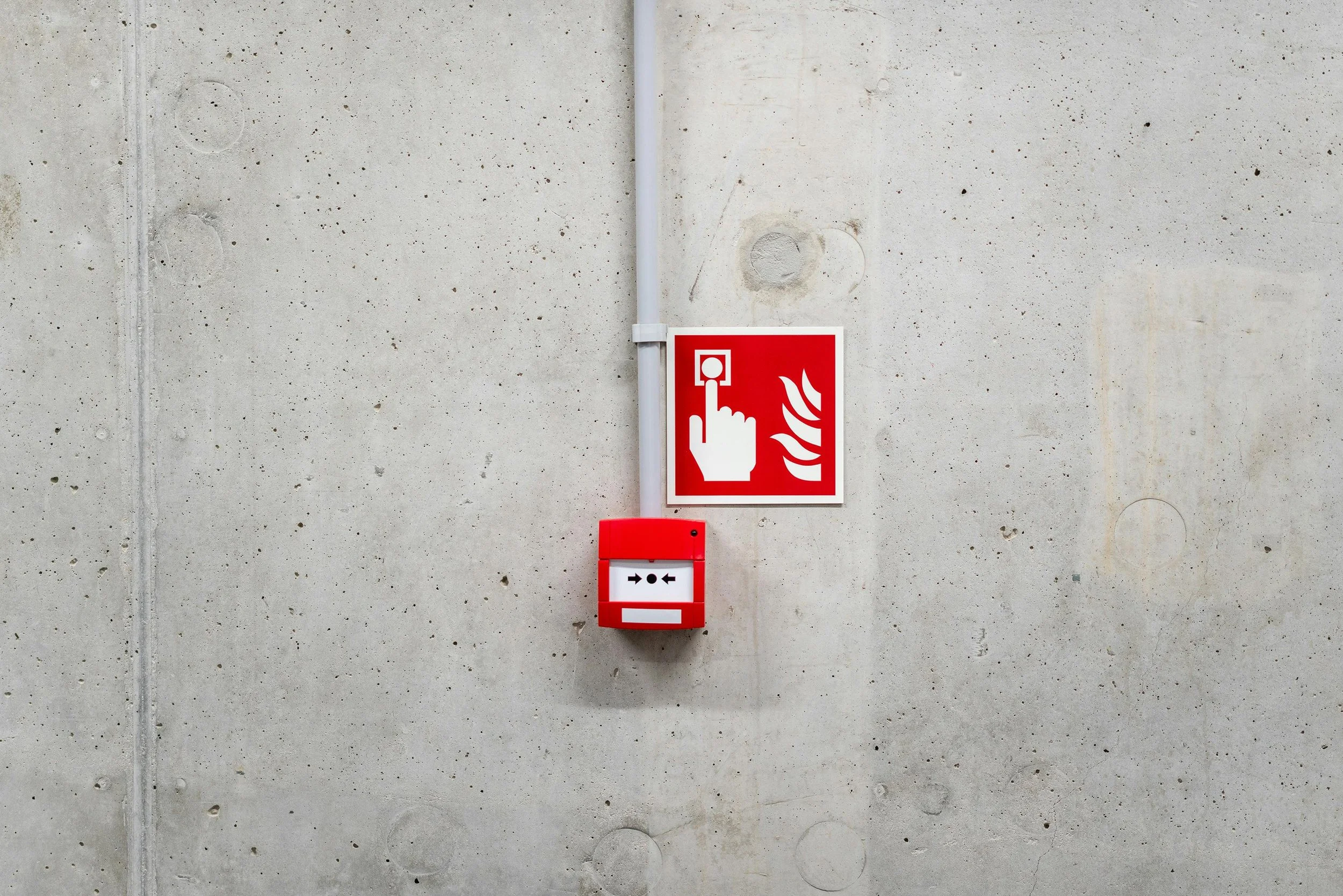 Red fire alarm box on a concrete wall with a sign above it depicting a hand pressing a button and flames.