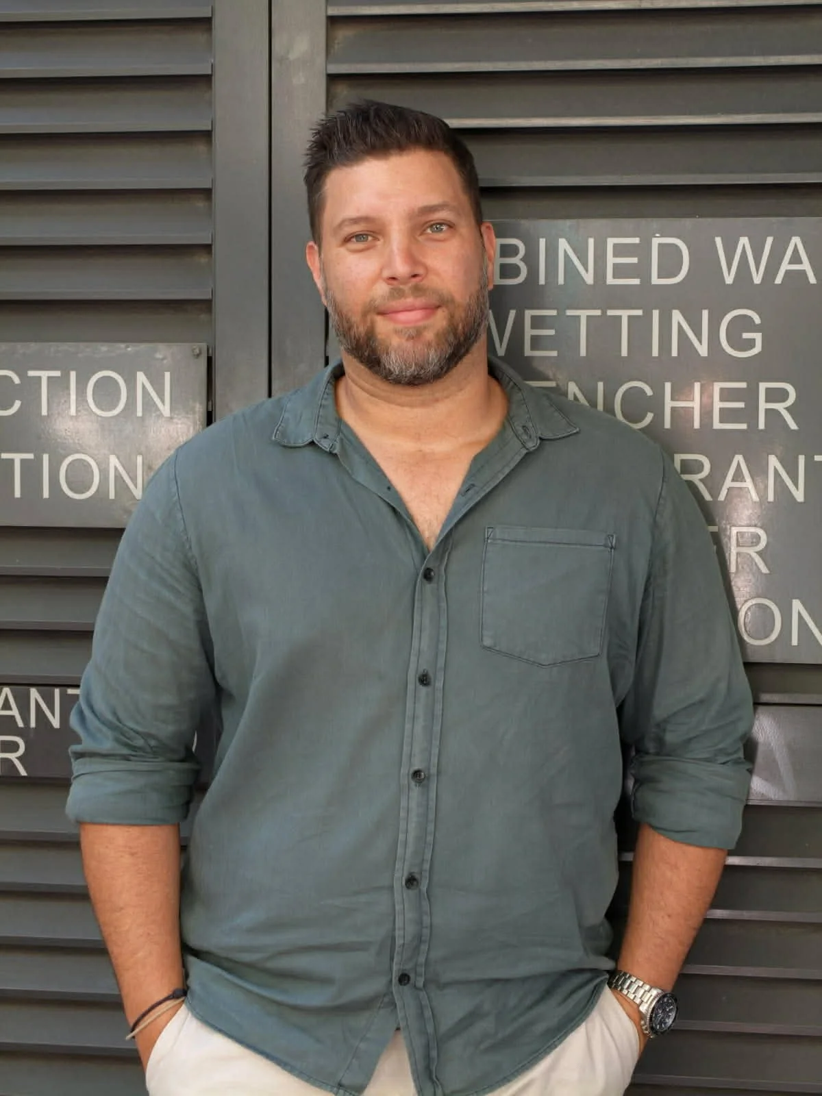 A man with a beard and short dark hair, wearing a grey button-up shirt with rolled-up sleeves, standing outdoors in front of a grey louvered wall with partial text sign.