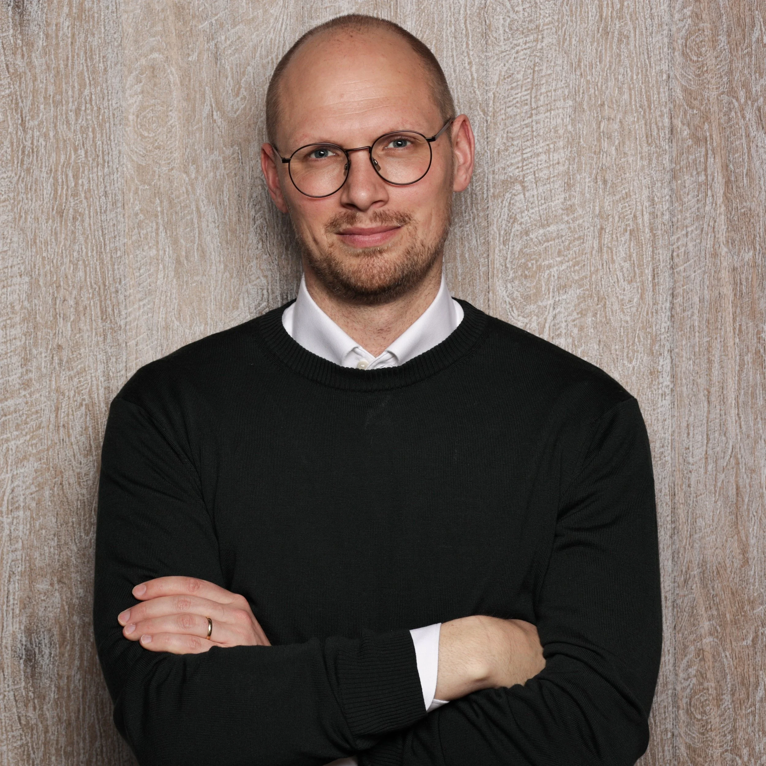 Laurenz Nienaber steht vor einer Holzwand und trägt eine schwarze Pullover mit weißen Hemd. Er hat kurze Haare, trägt runde Brillen und ist lächelnd.