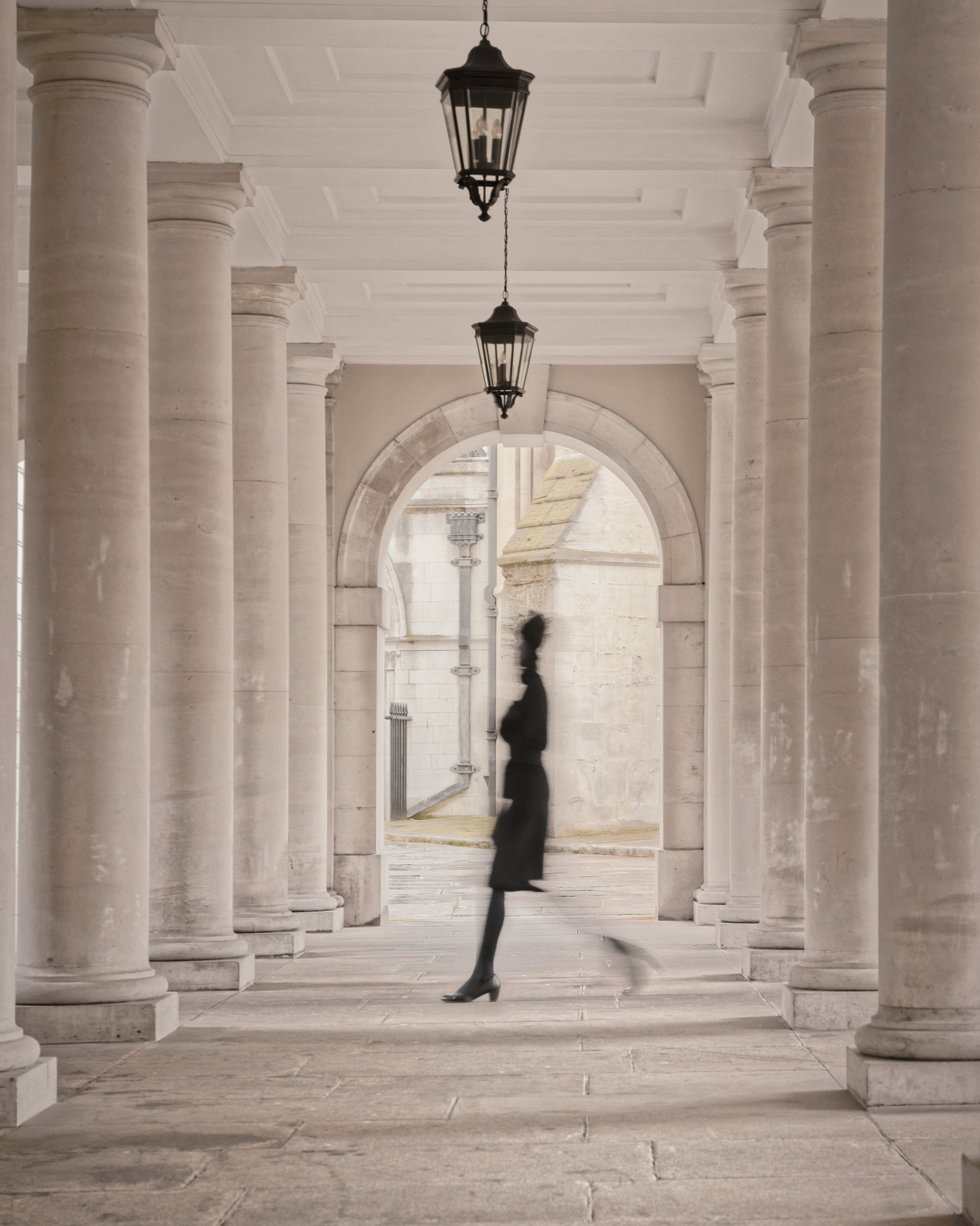 A person walking through a historical colonnade with large stone columns and hanging lanterns.