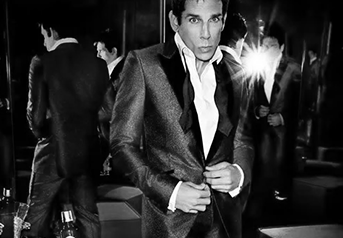 A man in a tuxedo adjusting his jacket at a formal event, with other people in tuxedos in the background.