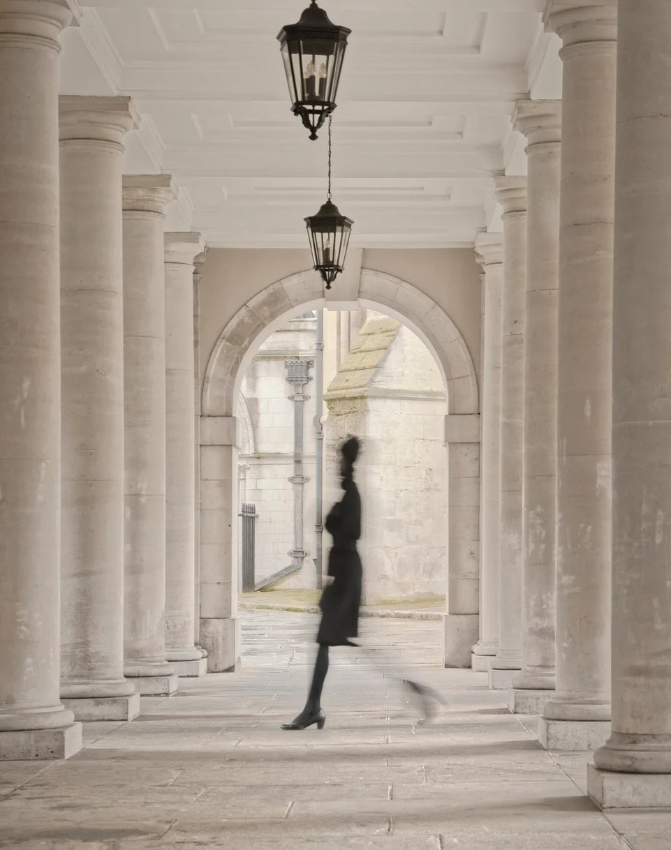 A silhouette of a woman in a long dress and high heels walking through a colonnade with stone columns and arches, with lanterns hanging from the ceiling.
