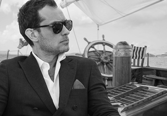 A man in a suit and sunglasses sitting outdoors, with a boat's steering wheel and deck in the background.