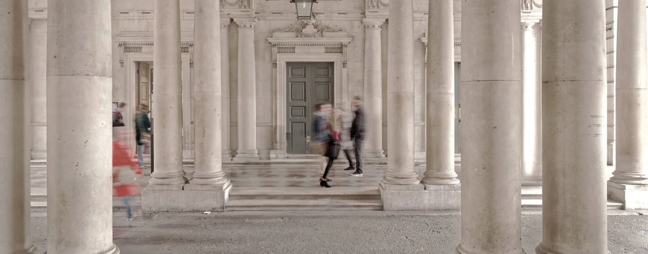 People walking in front of a historic stone building with columns and a dark wooden door