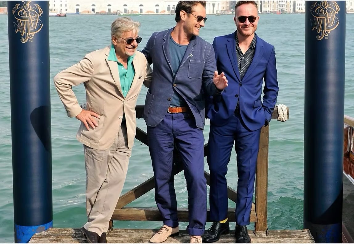 Three men in suits standing on a dock by the water, smiling and talking.