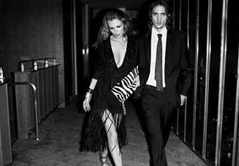 A man and woman walking together, dressed in formal black attire, in an indoor setting with metallic vertical bars in the background.