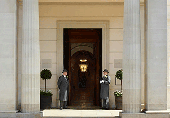 Two doormen standing outside a large building entrance with tall columns and a wooden door.
