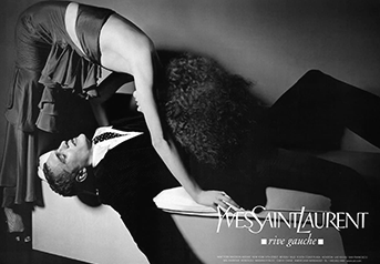 Black and white photo of a woman lying on her back on a table while another woman stands over her, leaning down and holding her arm. Text in the bottom right corner reads "Yves Saint Laurent".