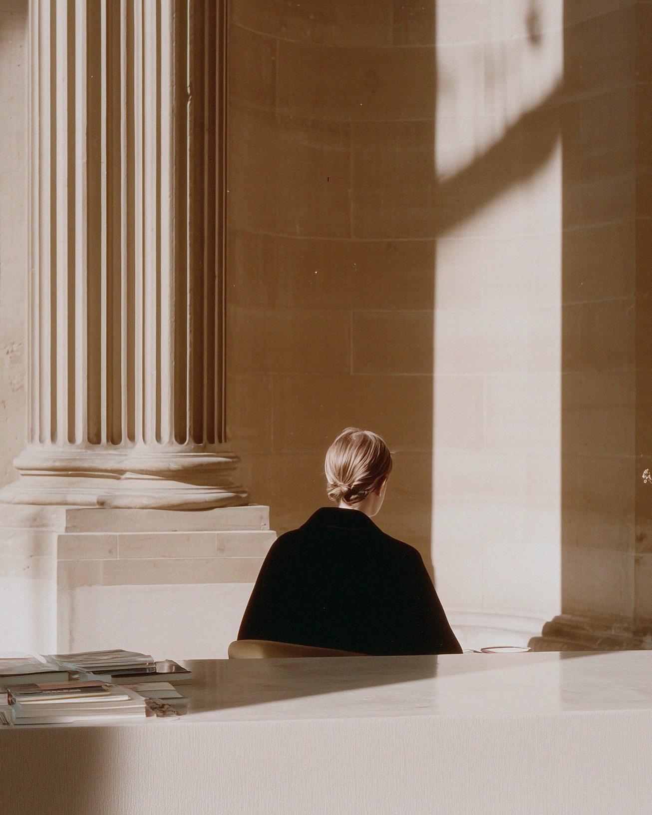 A person with blonde hair tied back, sitting at a desk in front of a large window with dark shadows and columns in a classical architectural style.