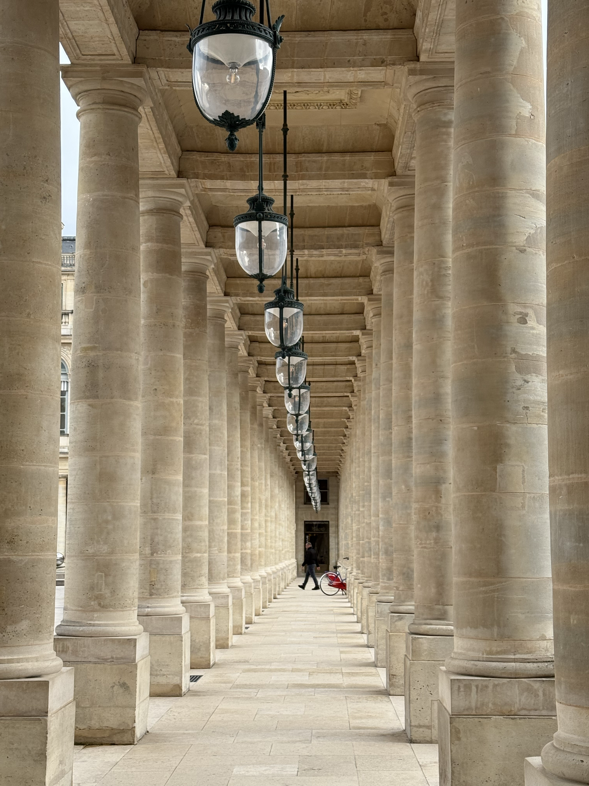 A covered walkway with tall stone columns and hanging black lanterns, with one person walking and pushing a bicycle with a child in a stroller.