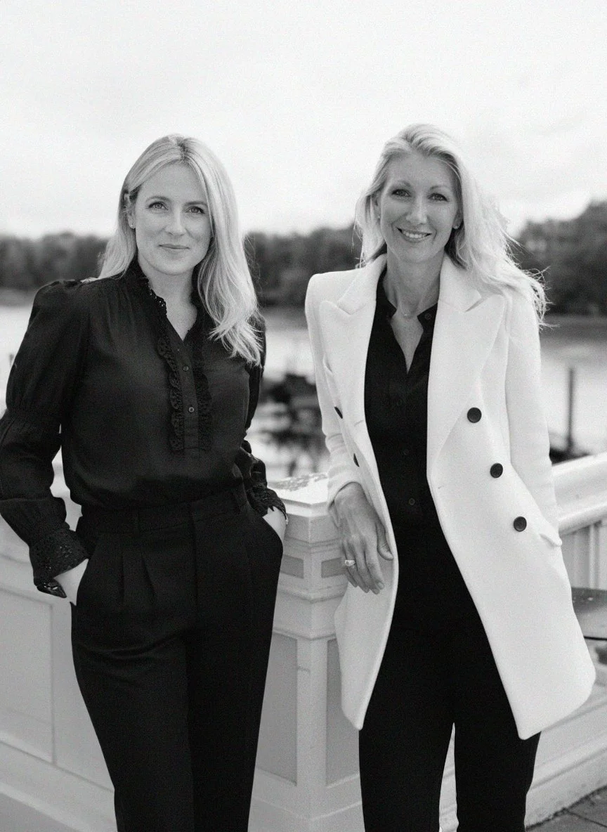 Two women standing outdoors near a body of water, dressed in black and white outfits, smiling at the camera.