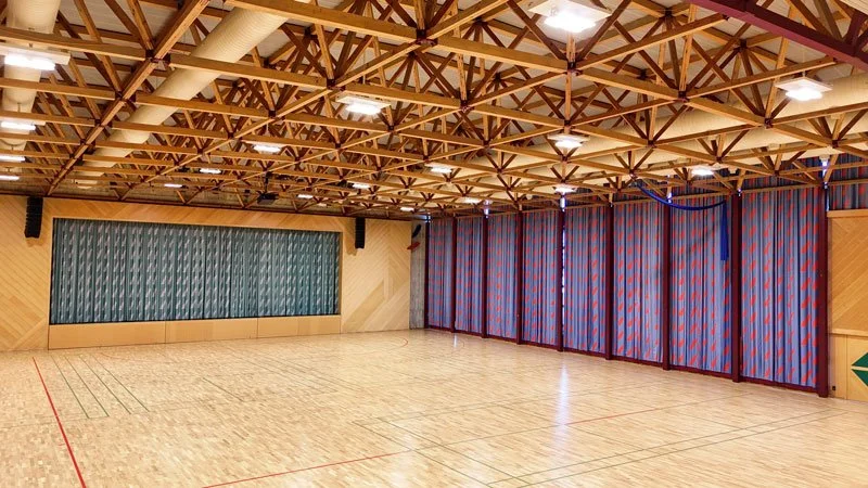 Leerer Turnraum mit Holzboden, grossen blauen Vorhängen und einem Holzbalkendecke.