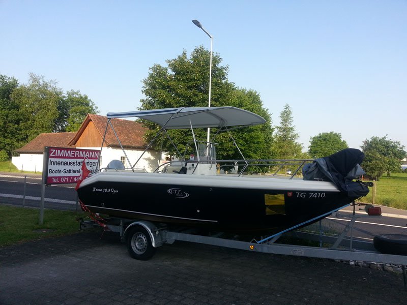 Ein Boot auf einem Anhänger, parkiert neben einer Strasse mit einem Haus und Bäumen im Hintergrund, sonniges Wetter.