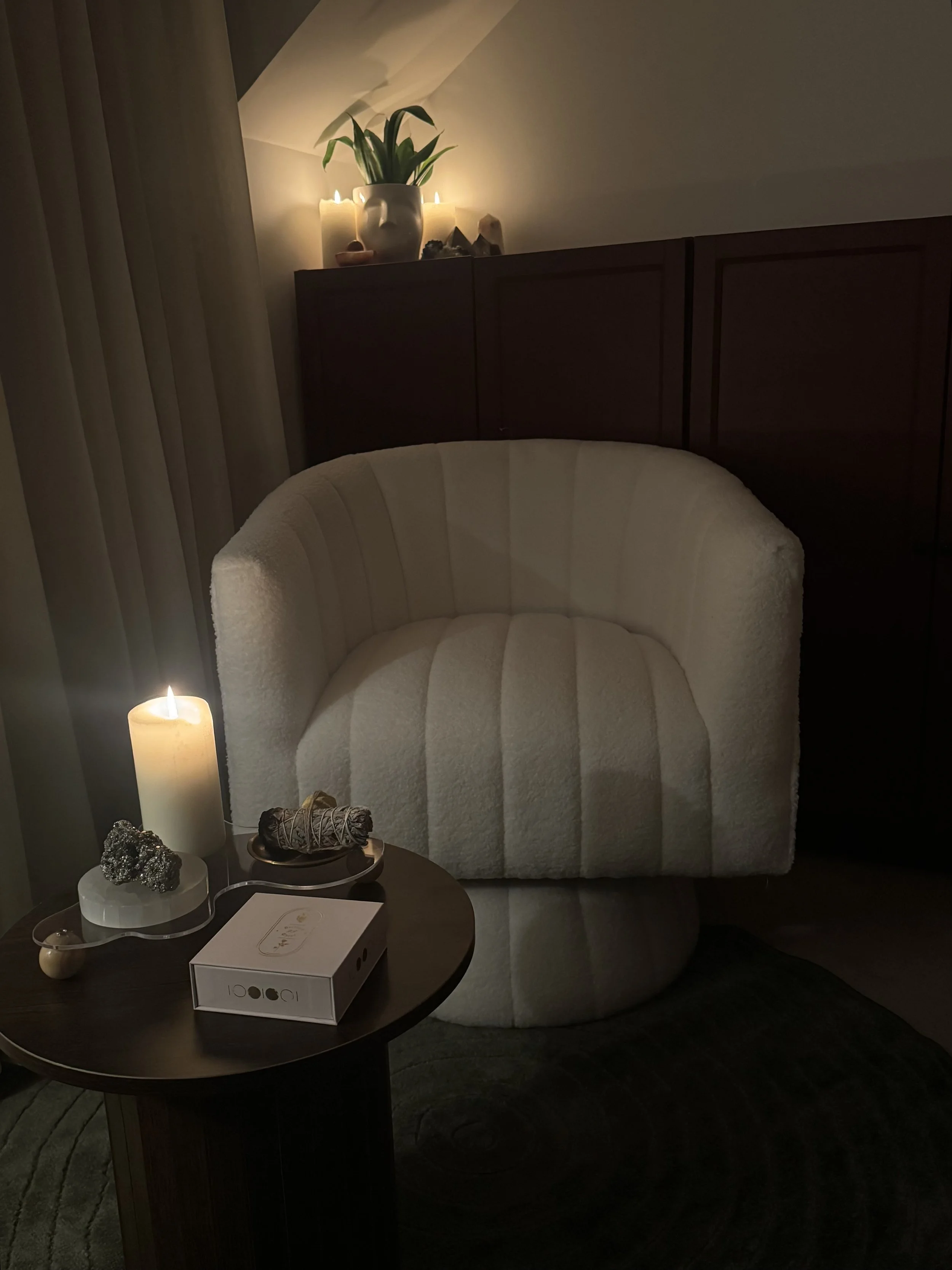 A cozy sitting area with a white, textured armchair, a round side table with candles and decorative items, and a dark wood cabinet behind the chair, topped with candles, a potted plant, and stones. Soft lighting and candles create a warm atmosphere.
