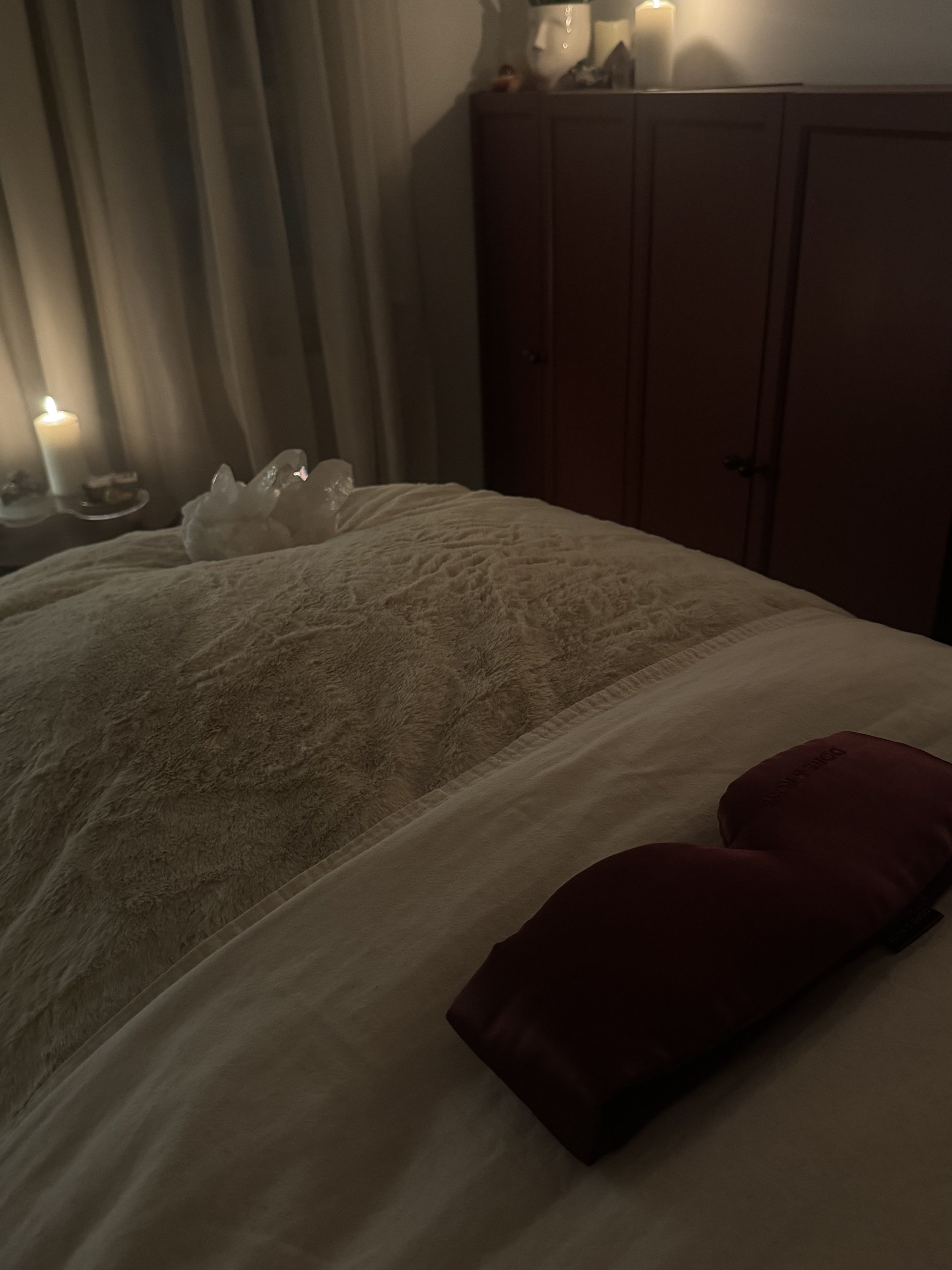 Dimly lit bedroom with a bed covered in a soft, beige blanket. On the bed is a Burgundy-colored heating pad. To the side, near the window, is a lit candle with a few white decorative stones in front of it. A dark wooden cabinet is in the background, and a charging or electronic device is visible on top of it.