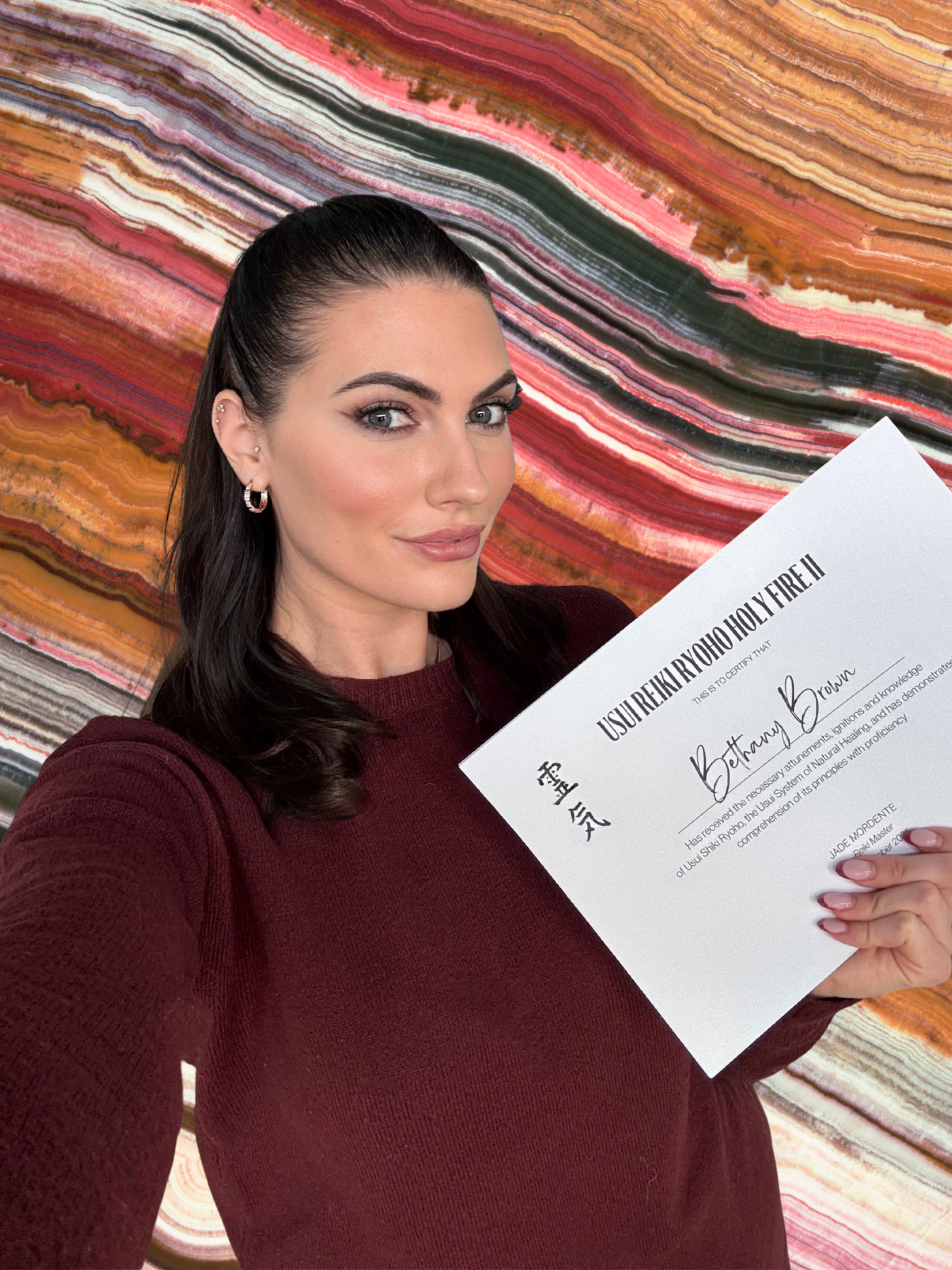 Soul Stories founder Bethany with dark hair, wearing a maroon sweater, holding a certificate of Reiki qualification in front of a colorful, striped background.