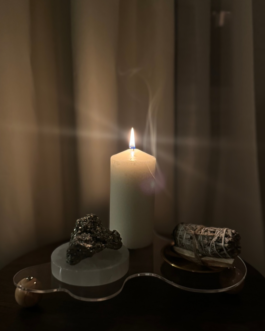 A lit candle on a table with a piece of black mineral, a bundle of sage tied with string, and a decorative glass tray.