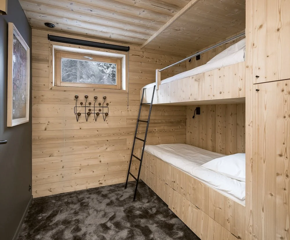 Chambre avec lit superposé en bois, petite fenêtre avec vue sur la neige, mur en bois, tapis gris, porte noire et crochets muraux.