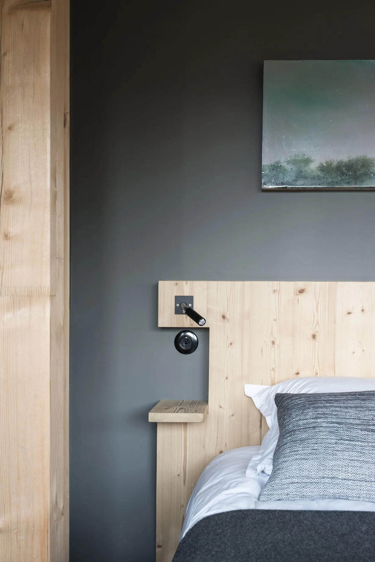 Coin de chambre avec lit en bois clair, lampe de lecture noire, et tableau abstrait au mur gris.