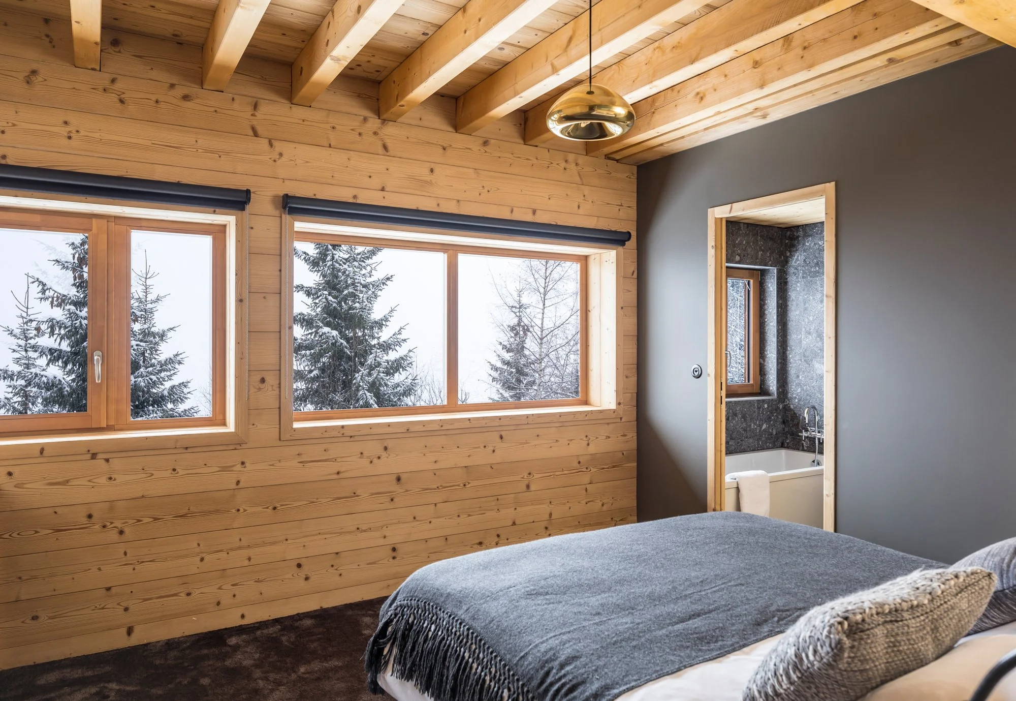Chambre avec murs en bois, fenêtres vers une forêt enneigée, lit avec couverture gris foncé, porte donnant sur une salle de bain avec baignoire et fenêtre, plafond en bois avec luminaire en métal doré.