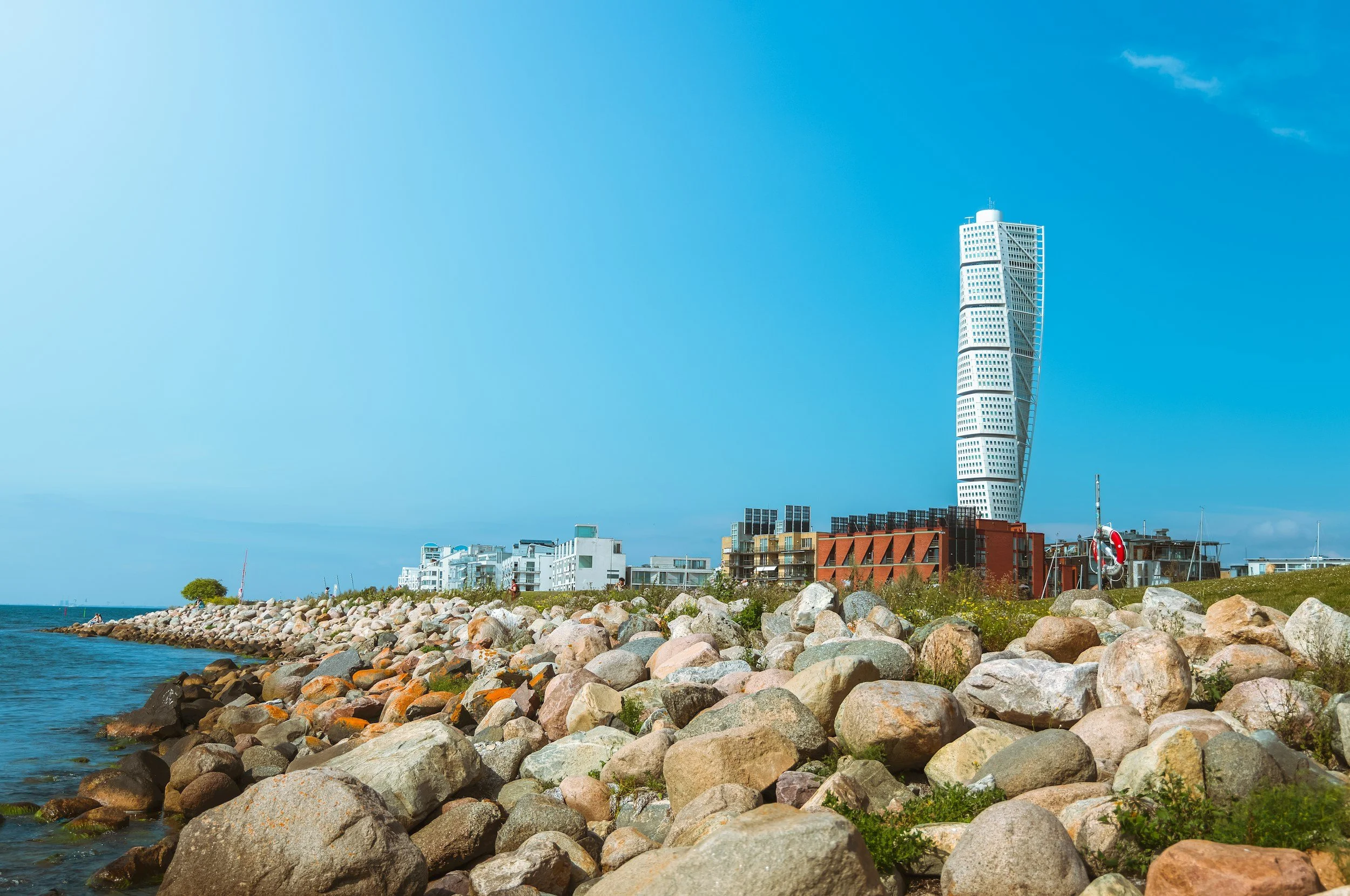 A cityscape with modern high-rise buildings next to a rocky waterfront and a clear blue sky.