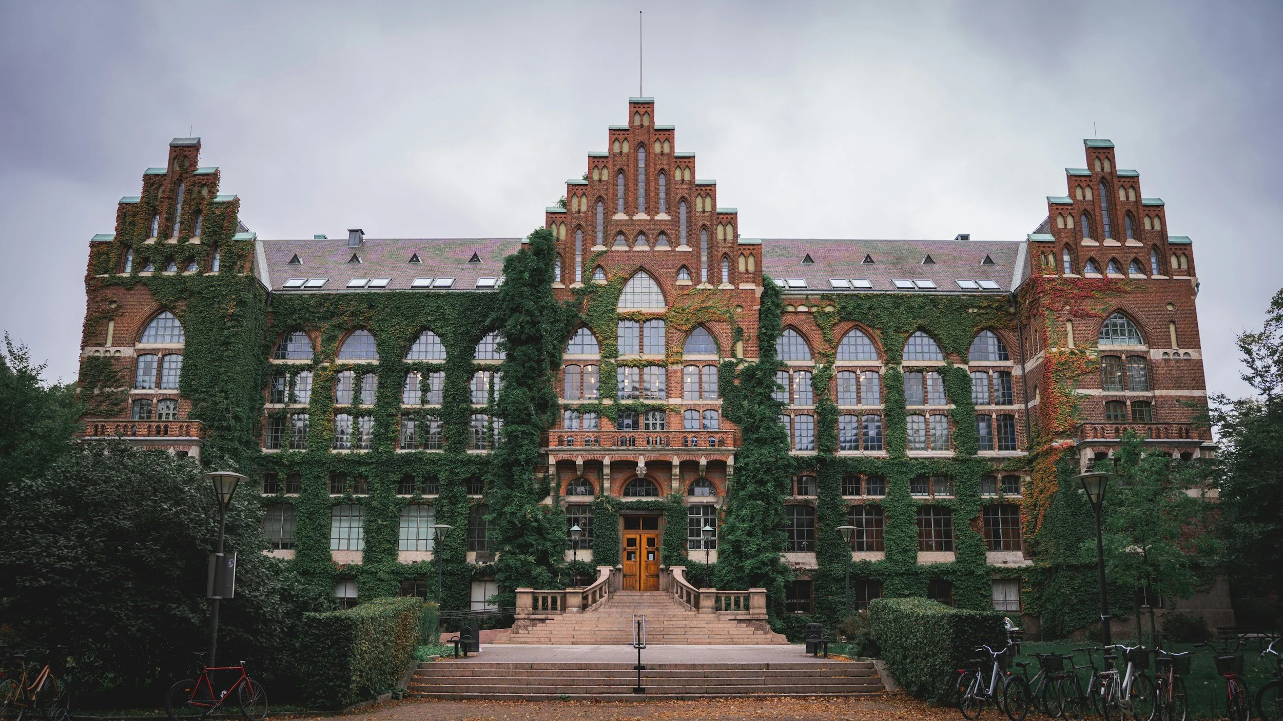 Facade of a large, historic brick building covered partially with green ivy, featuring tall arched windows, decorative brickwork, and a central tower with stepped gables, set against a cloudy sky.