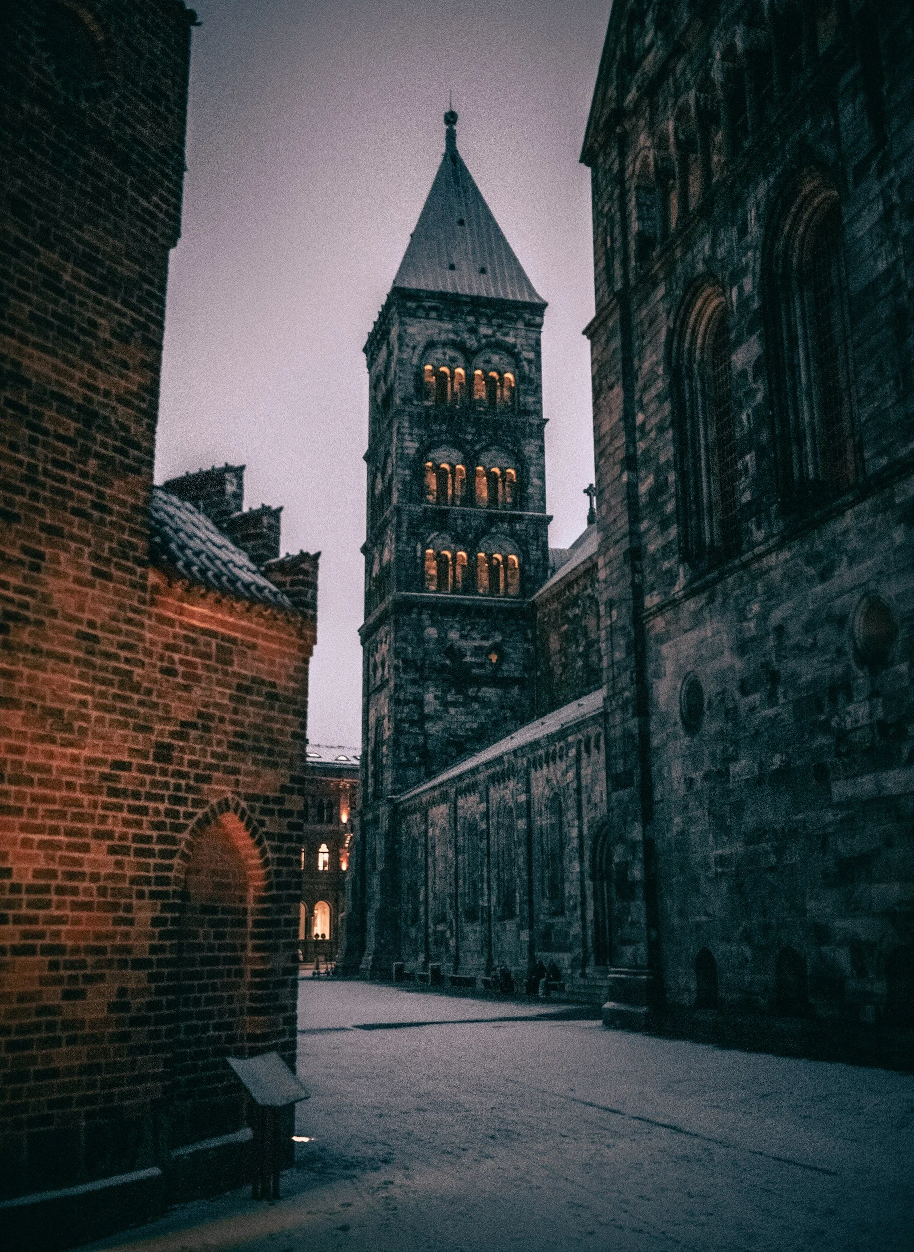 Historic stone church tower with illuminated arched windows between medieval brick and stone buildings at dusk