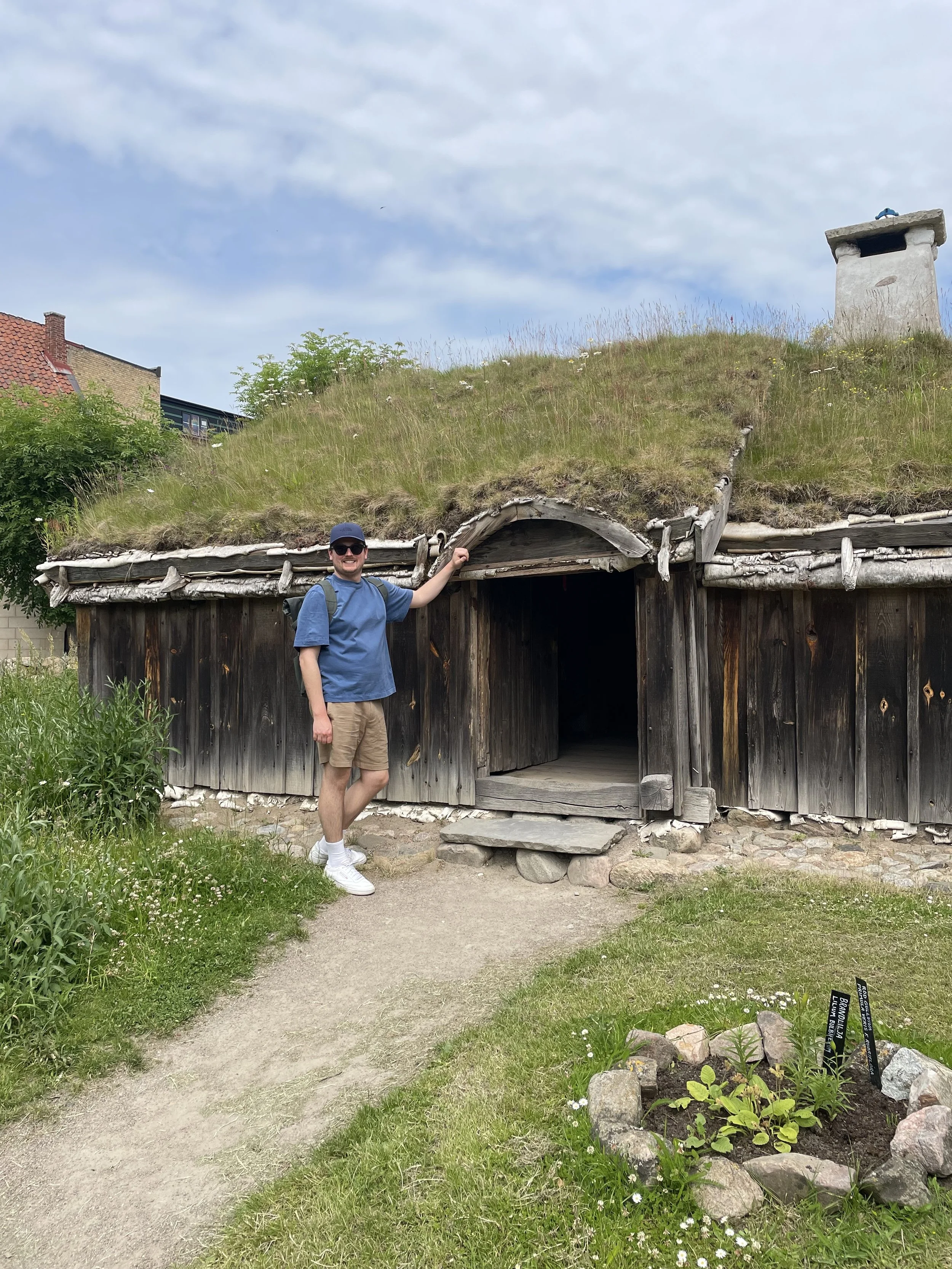 A man wearing sunglasses, a blue t-shirt, tan shorts, and a gray cap standing in front of a small wooden hut with a grass-covered roof, smiling and pointing at the hut's entrance. There is a pathway and a flower bed with small white flowers and stones nearby, under a partly cloudy sky.