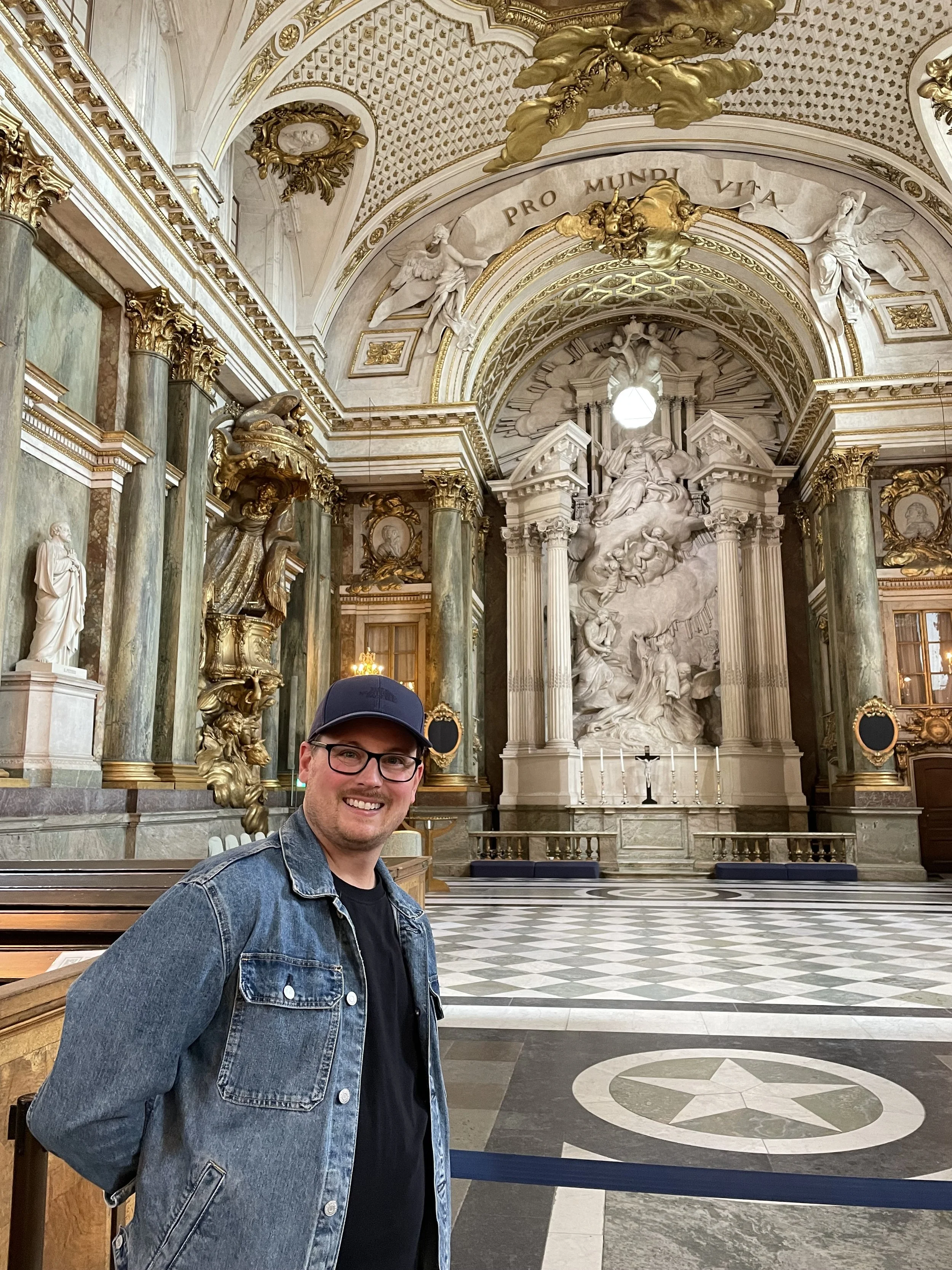 A smiling man wearing glasses, a black cap, and a denim jacket standing inside a grand, ornate church with gold accents, marble columns, and a large intricate altar with sculptures and columns.