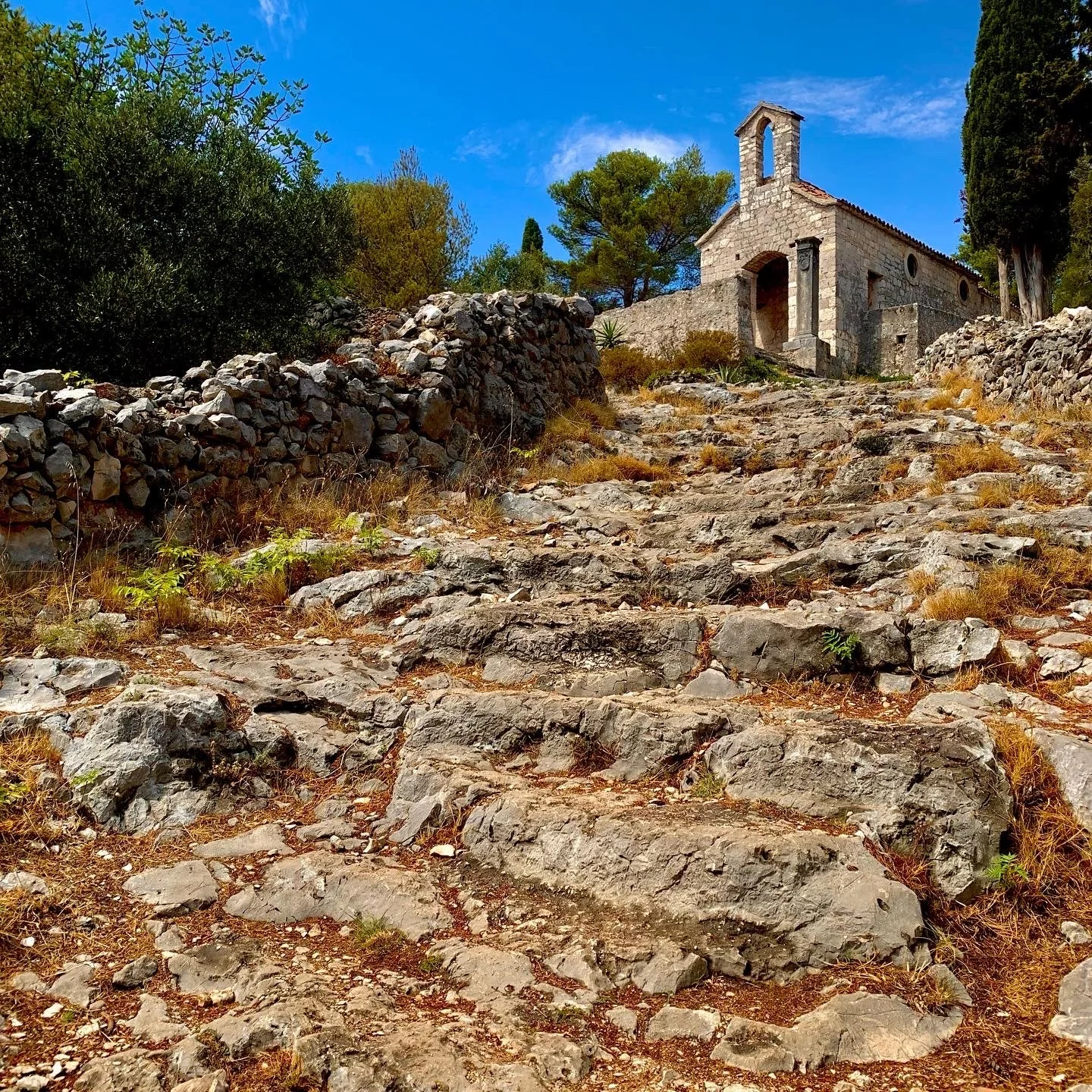 Stone pathway or stairs leading up to an old stone church or chapel on a hillside, surrounded by trees and greenery, under a bright blue sky.