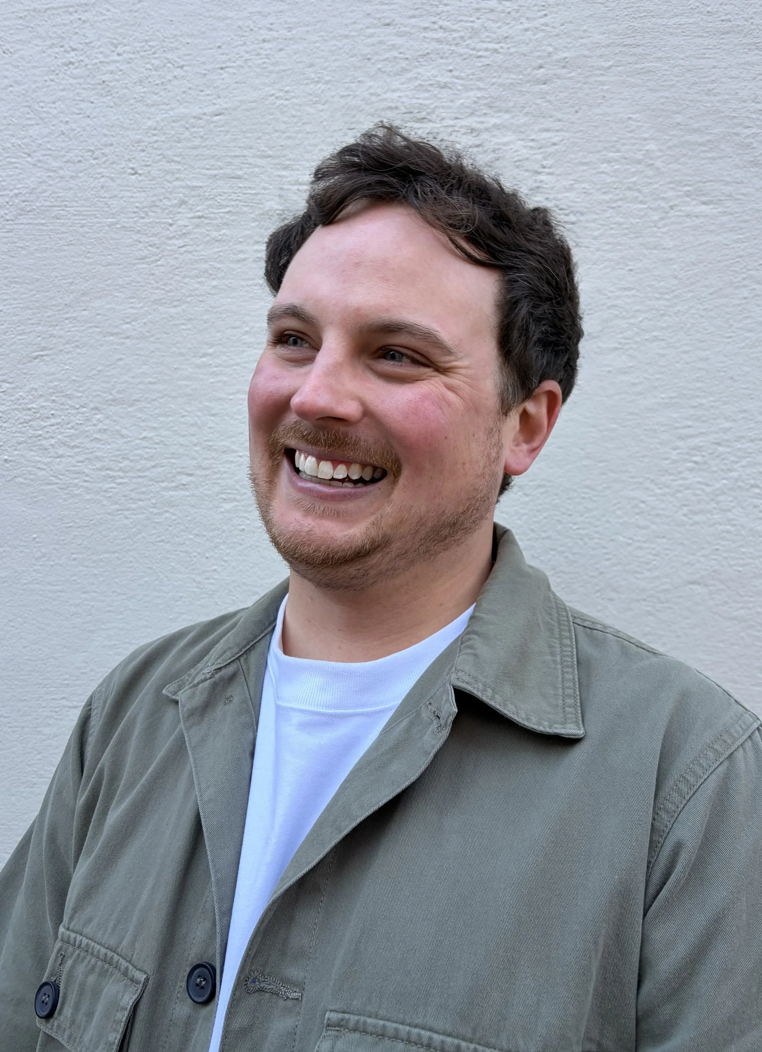 A smiling man with short dark hair and a beard, wearing a light green jacket over a white t-shirt, standing against a plain light-colored wall.