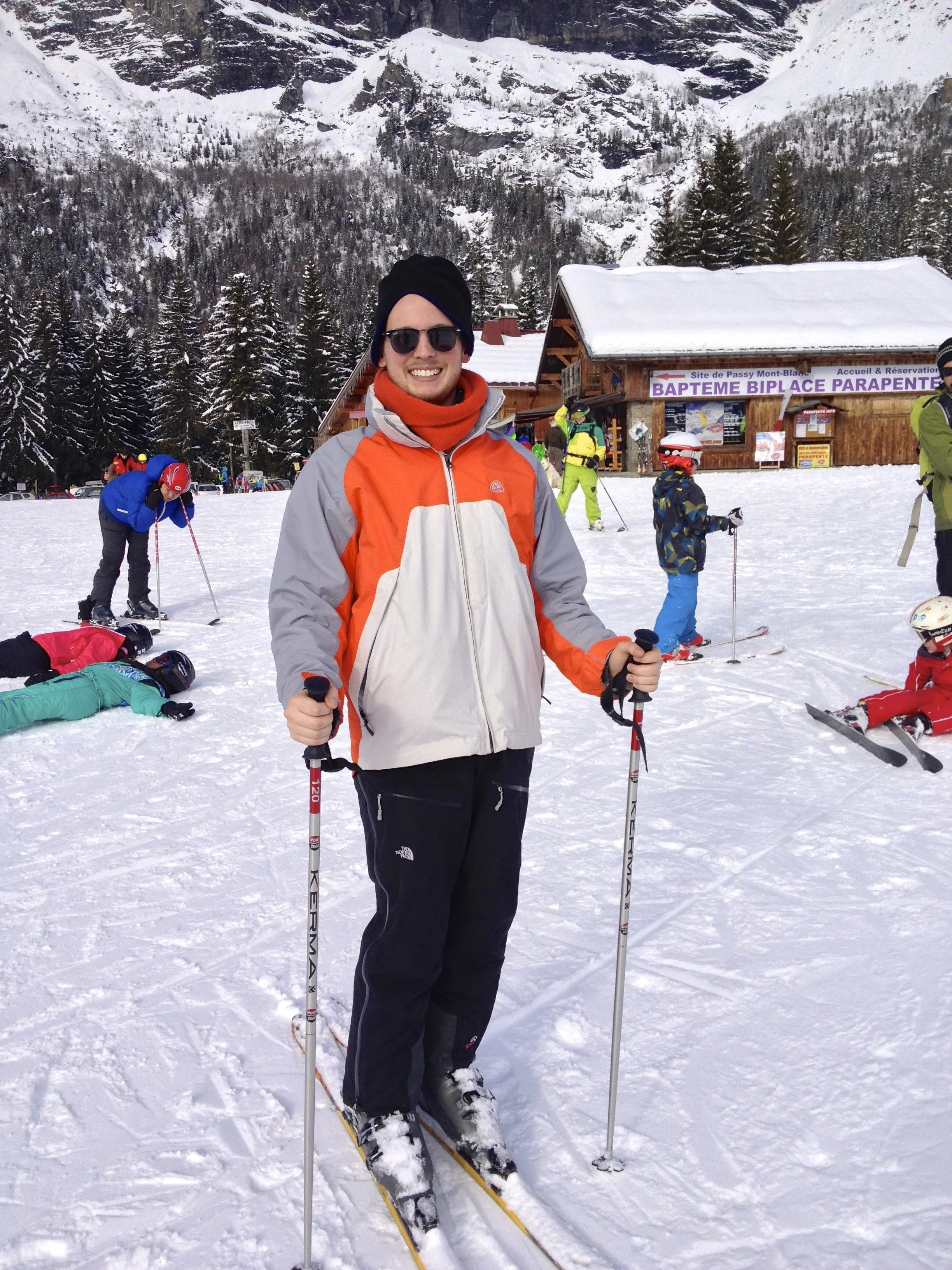 A person smiling in skiing gear holding ski poles on snow at a ski resort, with a wooden building and other children and adults skiing in the background, snow-covered trees and mountains.