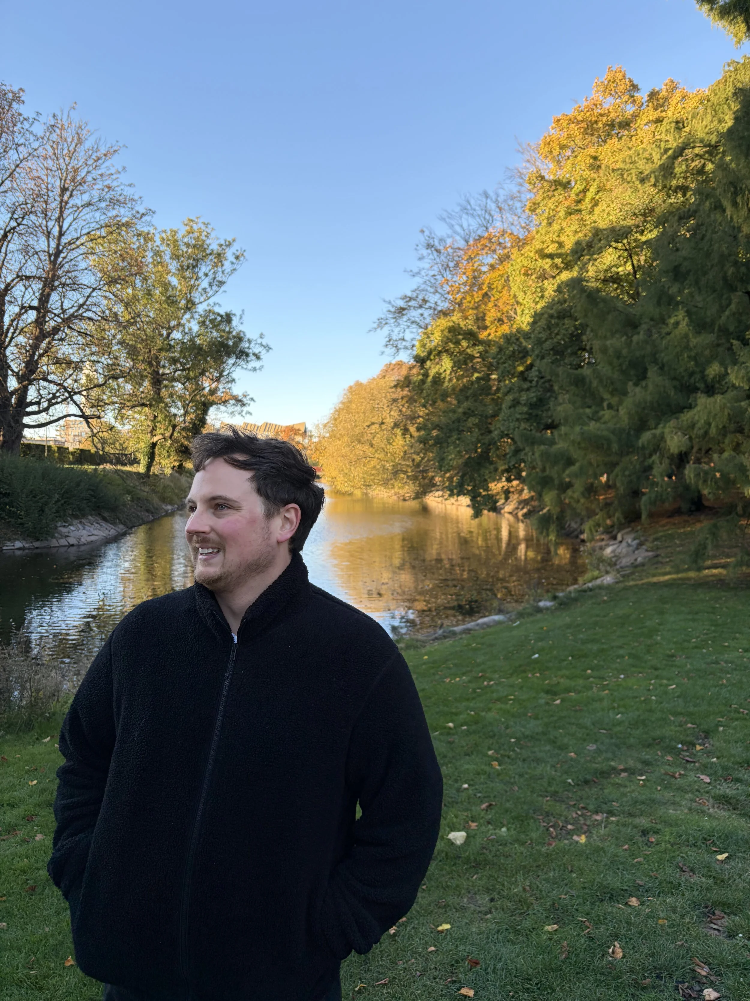 A young man wearing a black fleece jacket standing outdoors near a waterway with trees exhibiting fall foliage, under a clear blue sky.